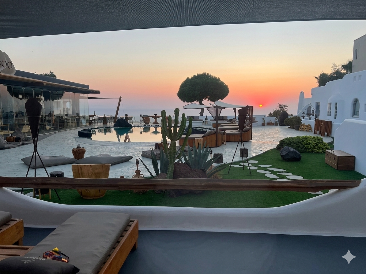 Sunset view of a terrace with lounge chairs, a small pool, and decorative plants, overlooking the ocean with a tree and buildings in the background.
