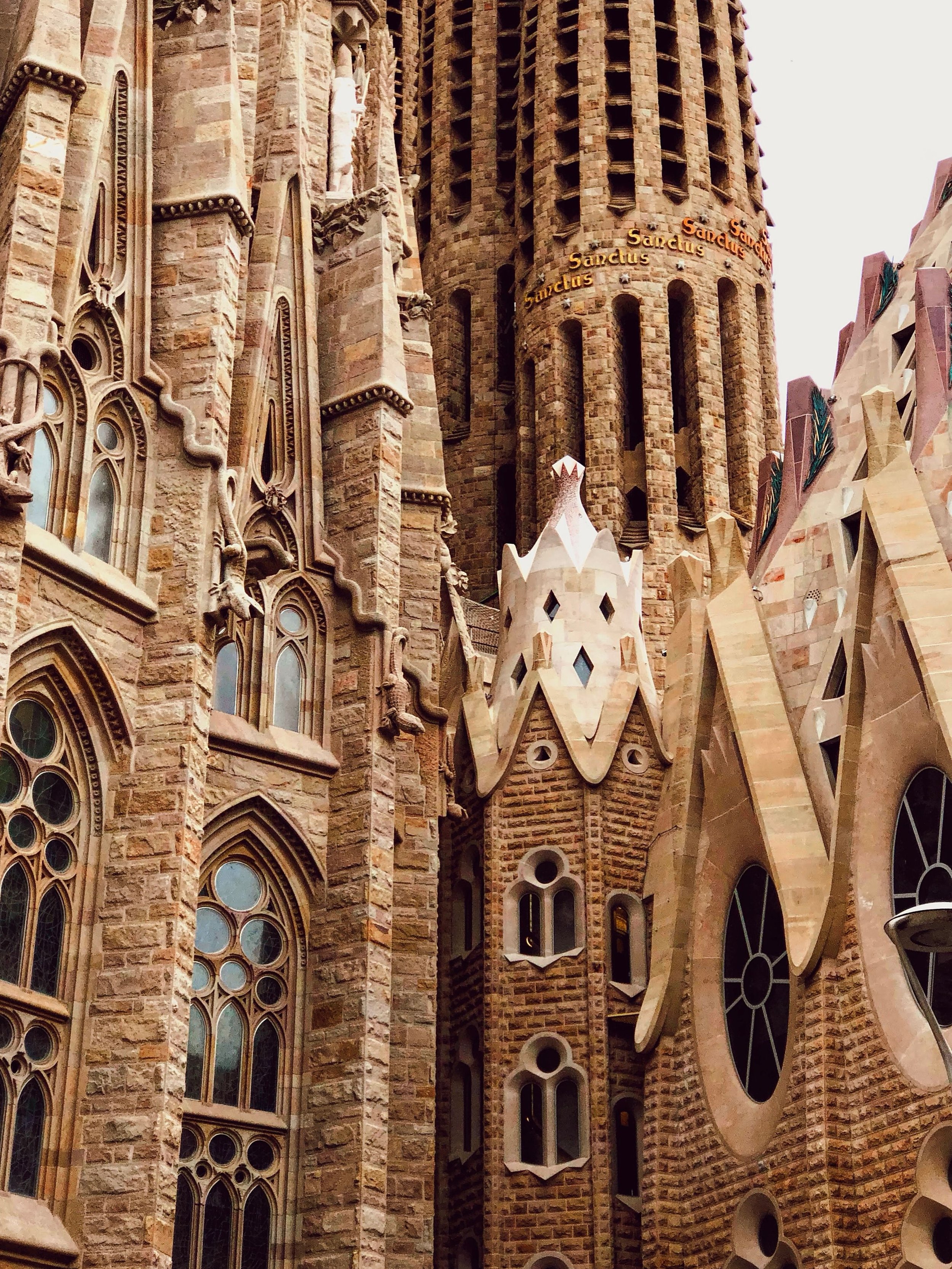 Close-up of the Sagrada Familia's intricate stone architecture, showing tall towers, colorful stained glass windows, and detailed sculptures. The word 'Sanctus' is visible on the tower.