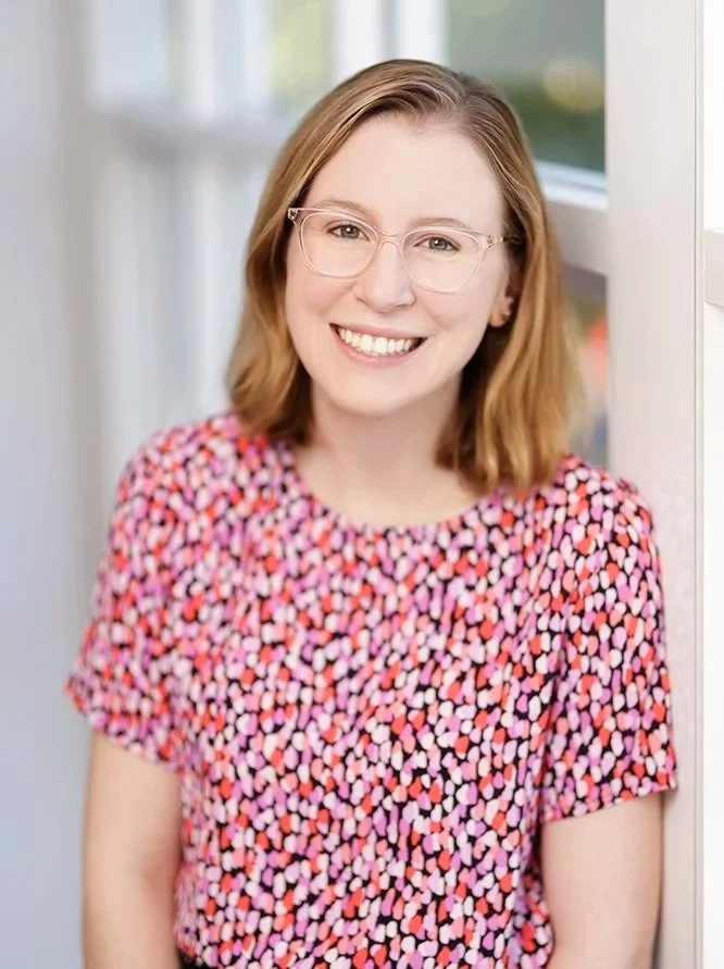 A young woman with shoulder-length reddish hair, wearing glasses and a colorful patterned shirt, smiling indoors near a window.