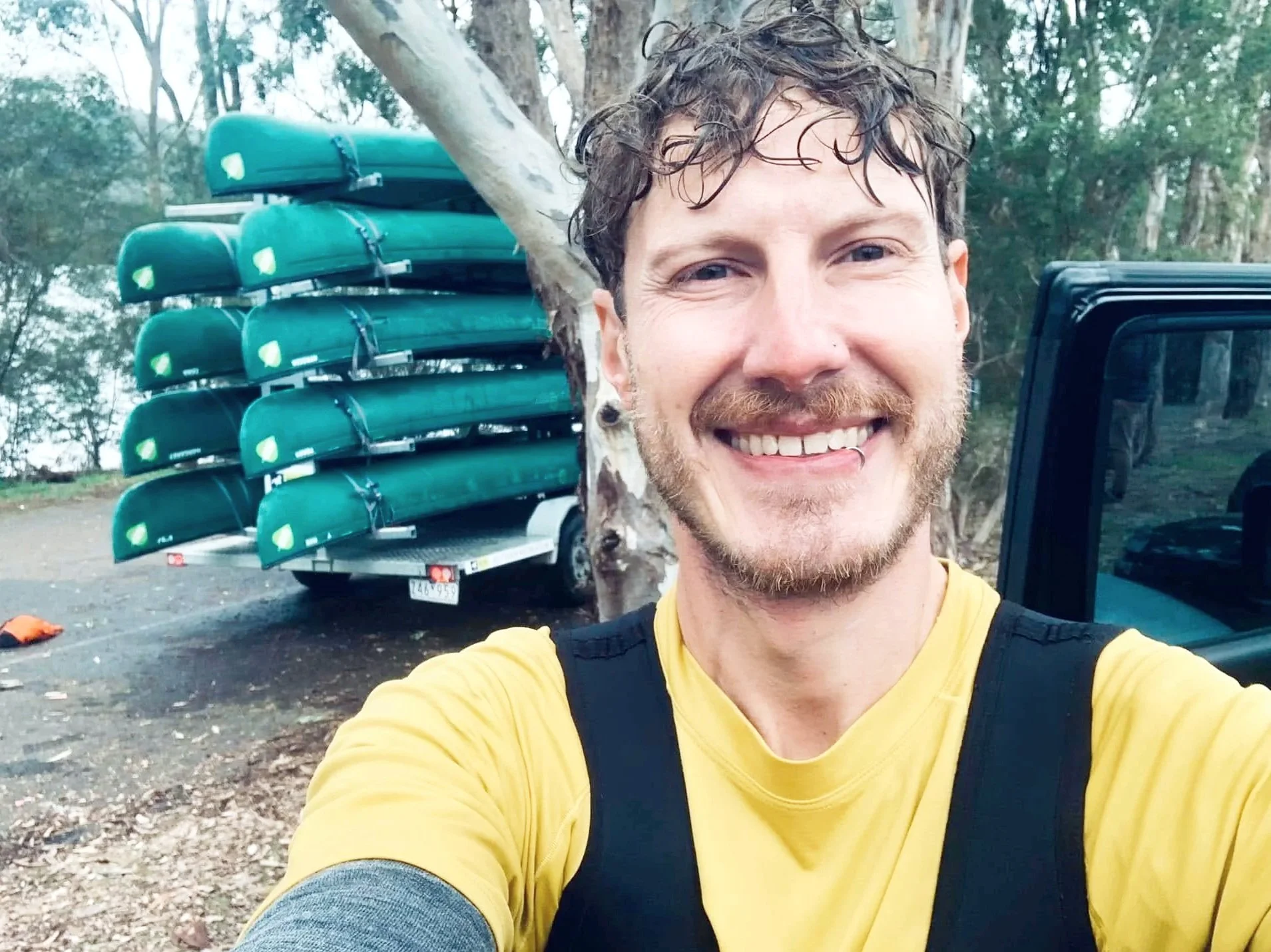 A smiling man with curly hair and a beard taking a selfie outdoors next to a tree. Behind him, there is a trailer loaded with green canoes and a jeep.