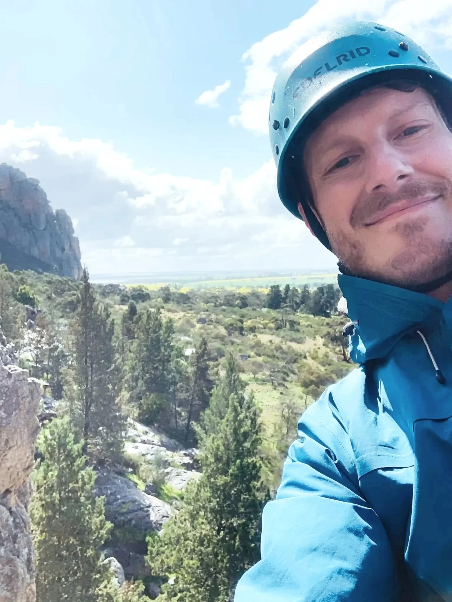 Close-up of a smiling man wearing a helmet and blue jacket, with a scenic outdoor landscape including rocky terrain, trees, and a distant plain under a partly cloudy sky.