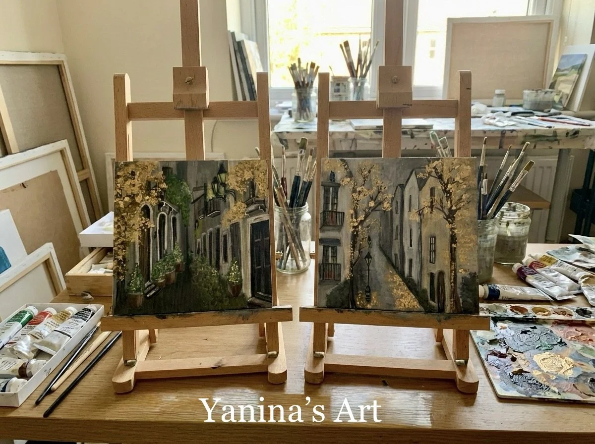 Two small canvases displaying finished landscape paintings of street scenes with trees and buildings.
