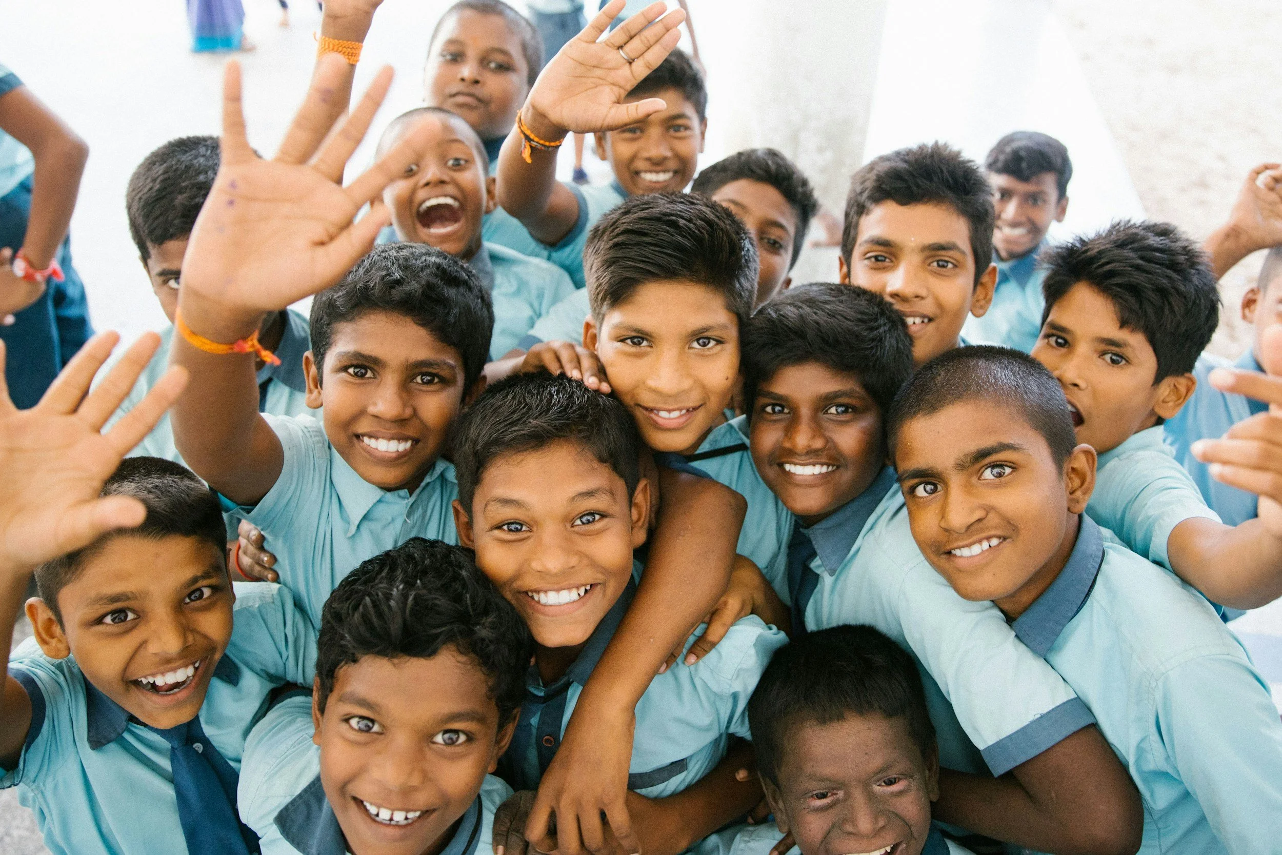 A diverse group of children wearing blue shirts, smiling and waving at the camera, some tightly grouped together.