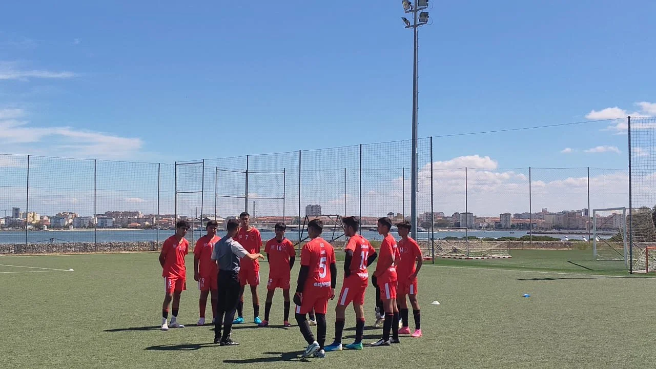 A youth soccer team in red uniforms listening to their coach during a practice session on an outdoor field with a cityscape and water in the background.