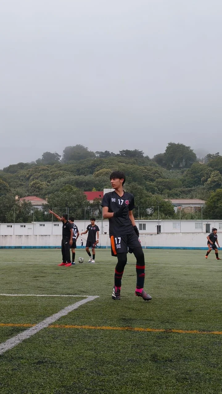 A group of soccer players on a field, with one player in the foreground stretching and others preparing for play in the background, surrounded by trees and a cloudy sky.
