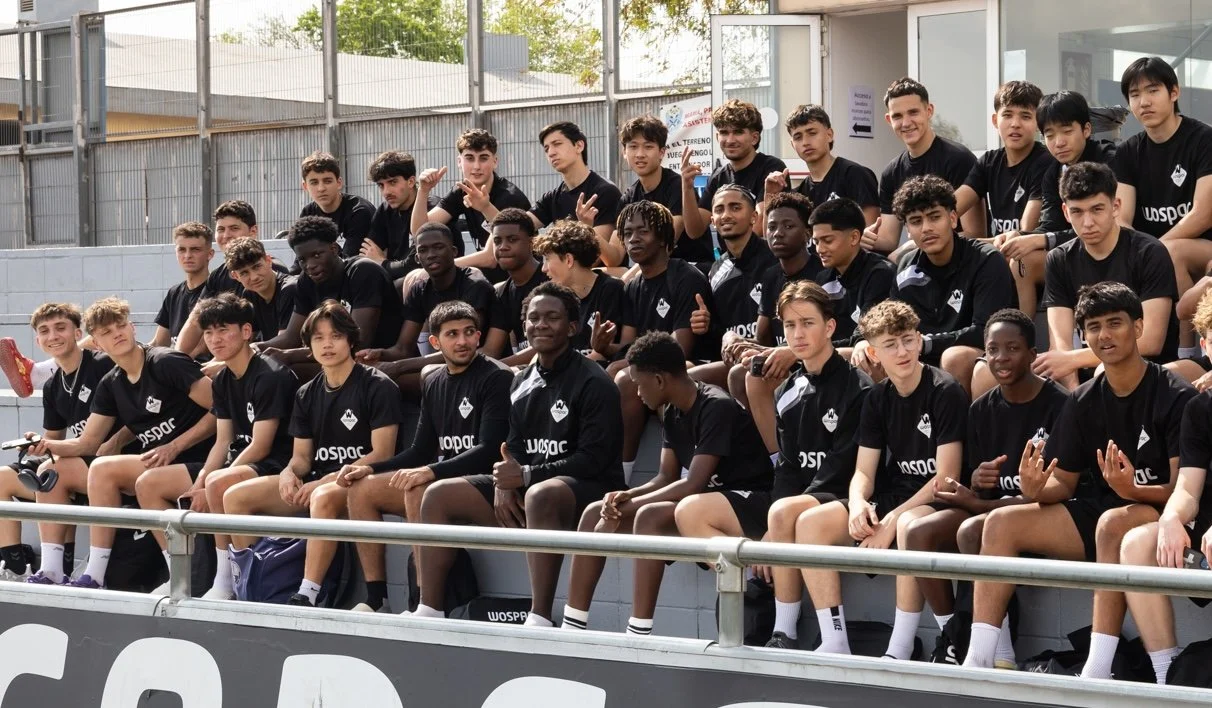 Group of young soccer players sitting on stadium bleachers outside, wearing matching black uniforms with 'WOSPAQ' logo.