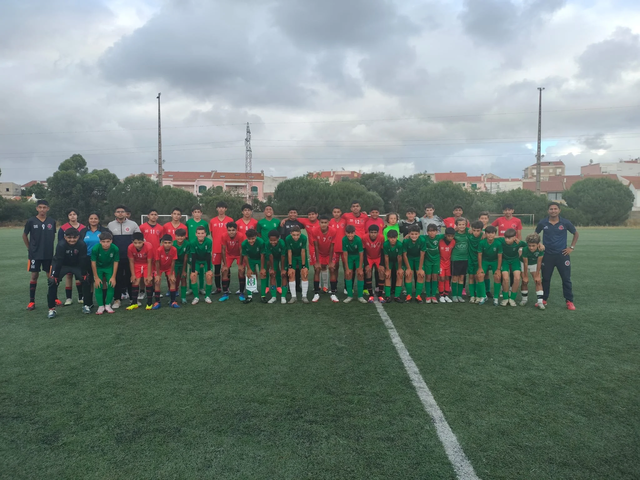 A large group of young soccer players and two coaches posing for a team photo on a soccer field. The players are dressed in red and green uniforms, standing and kneeling in front of a goal with trees and buildings in the background.