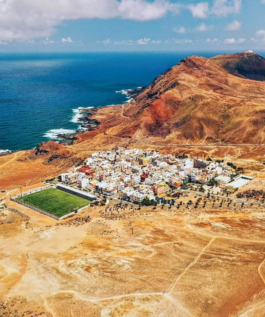Aerial view of a seaside town with a football field, tightly packed white buildings, mountainous terrain, and the ocean in the background.