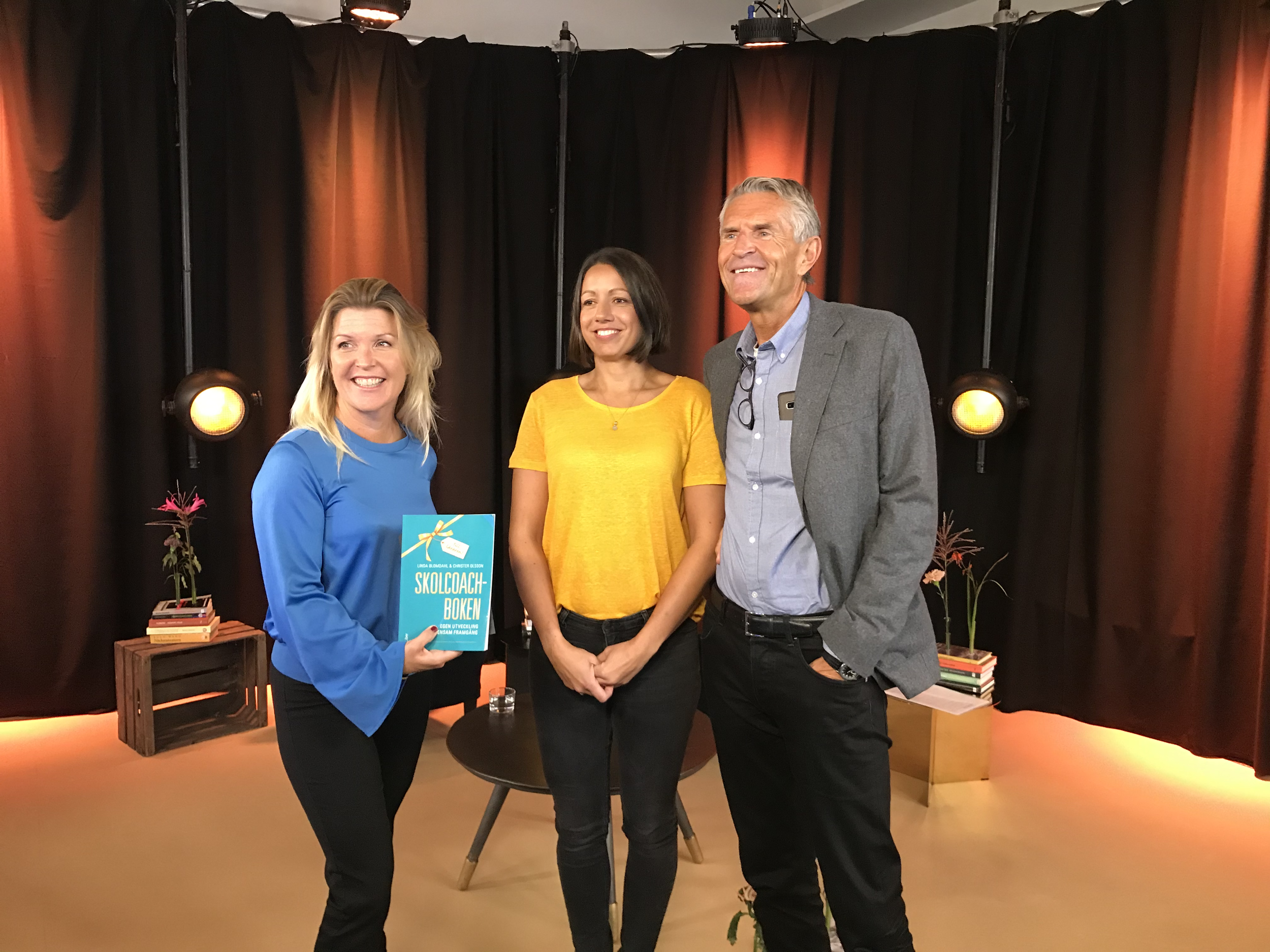 Three people standing together indoors, smiling for a photo. The woman in the middle wears a yellow shirt, the woman on the left wears a blue shirt and holds a book, and the man on the right wears a gray jacket and glasses hanging from his shirt. The background features brown and black curtains with stage lighting, and small book stacks and plants on tables.