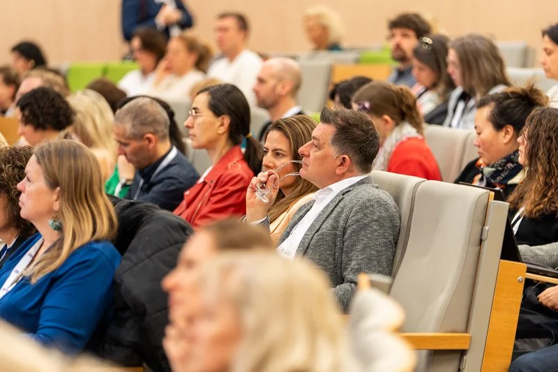 A diverse group of people attending a conference or seminar, sitting in tiered seats in a lecture hall.