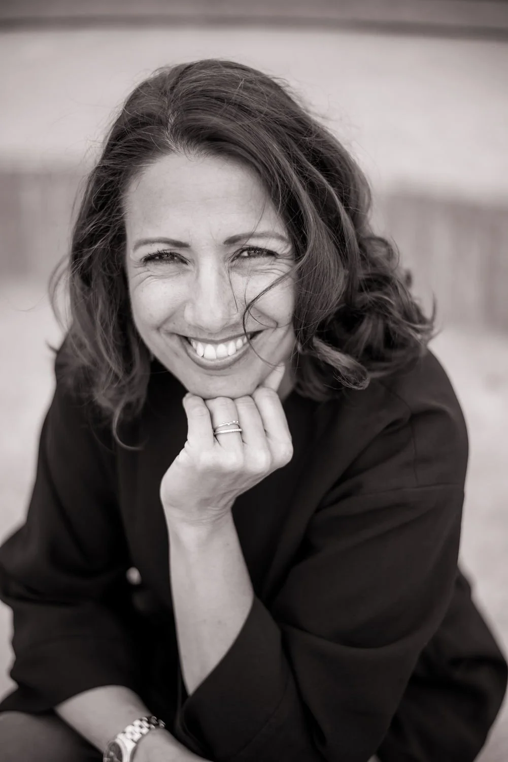 A woman smiling with her chin resting on her hand, wearing a dark top, in black and white photo.