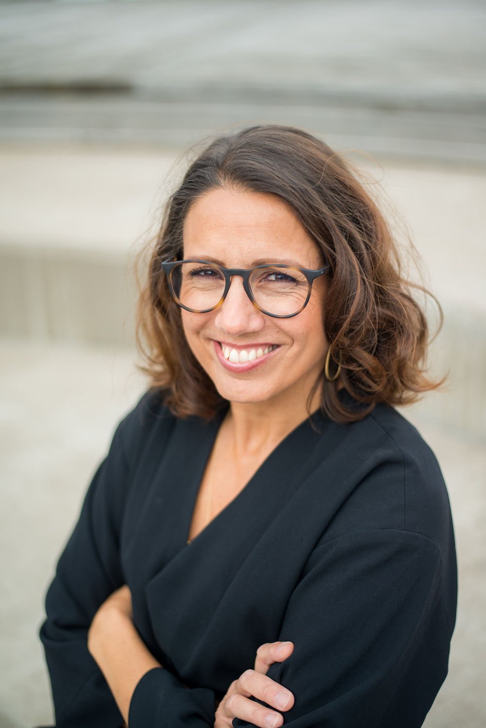 A woman with shoulder-length wavy brown hair and glasses, smiling and wearing a black blazer, standing outdoors with blurred background.