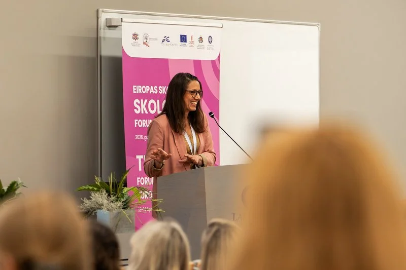 A woman with brown hair and glasses, dressed in a pink jacket, is standing at a podium giving a speech during a conference. There is a pink banner behind her with the text 'EUROPAS SKOLFORUM.' Audience members sit in front of her, and there are plants on the desks beside her.