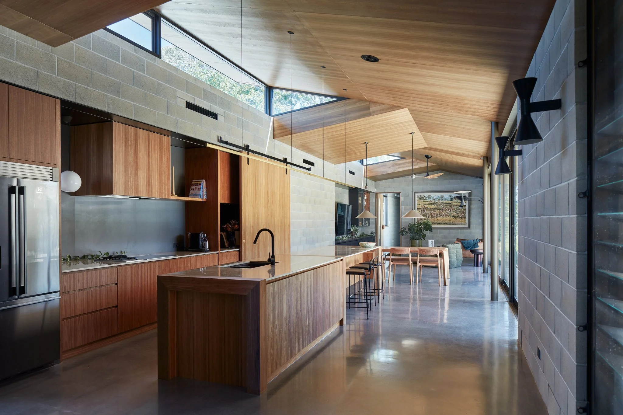 Modern open-concept kitchen with wooden cabinets, large island, stainless steel refrigerator, and a dining area with chairs. Features a high, angled wooden ceiling, large windows, and pendant lighting.