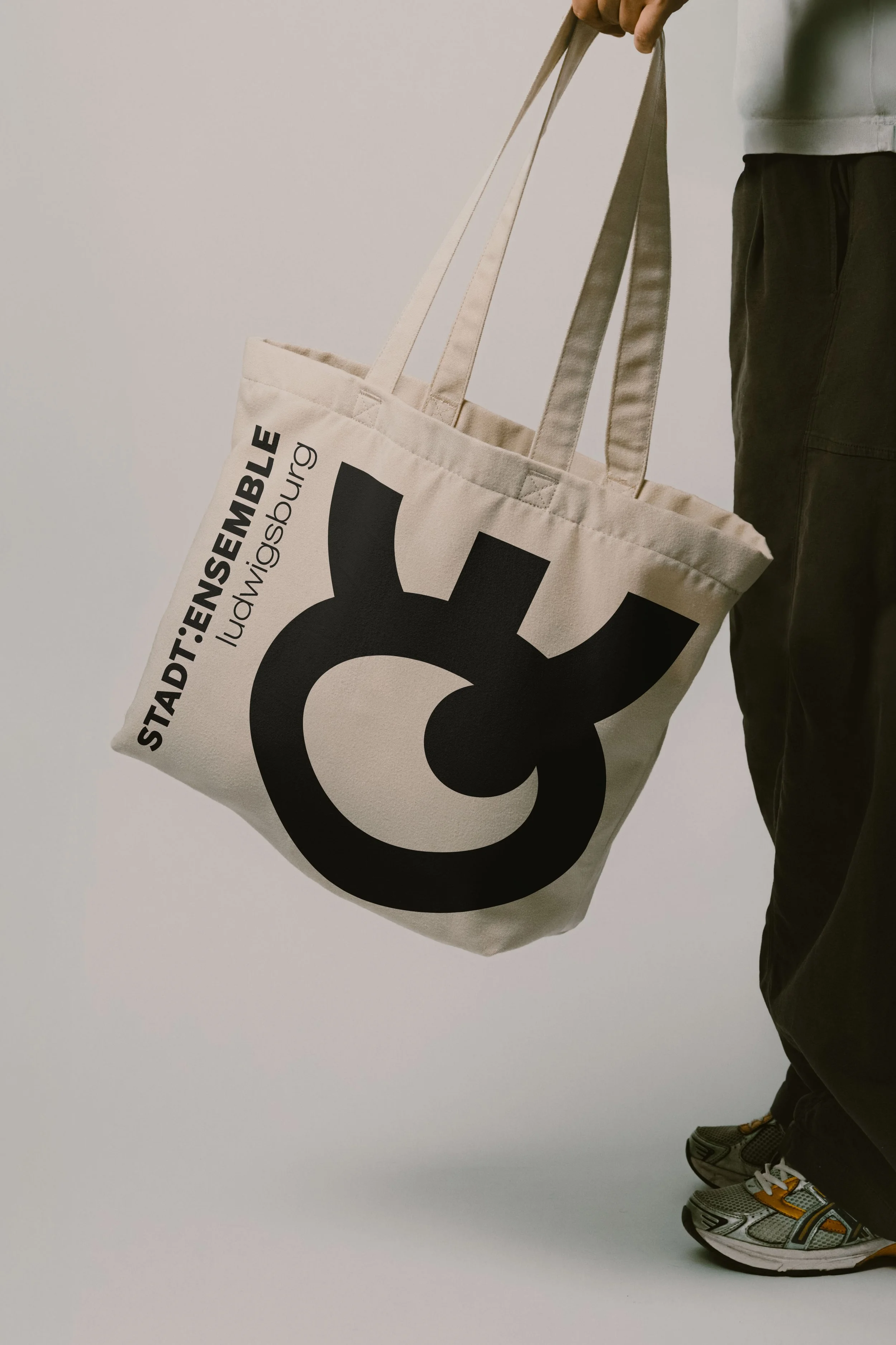 Person trägt eine weiße Stofftasche mit schwarzem Text und großem Logo, kleidet schwarze Hosen, steht auf bunten Sportschuhen.