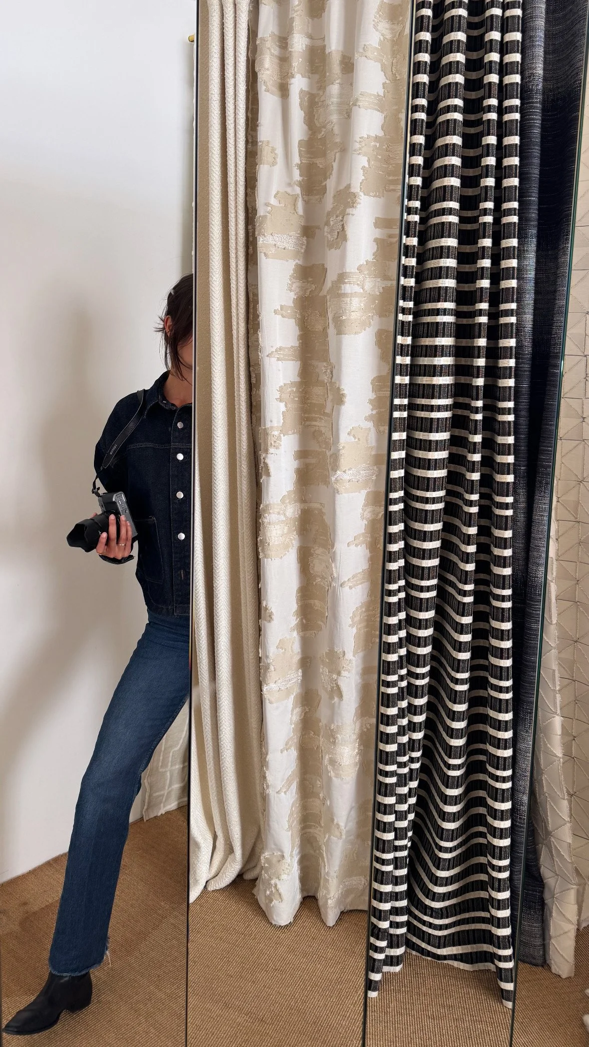 Eine Frau mit dunkler Jacke und blauer Jeans, die in einem Ankleidezimmer vor einem großen Spiegel steht, umgeben von verschiedenen Gardinenmustern