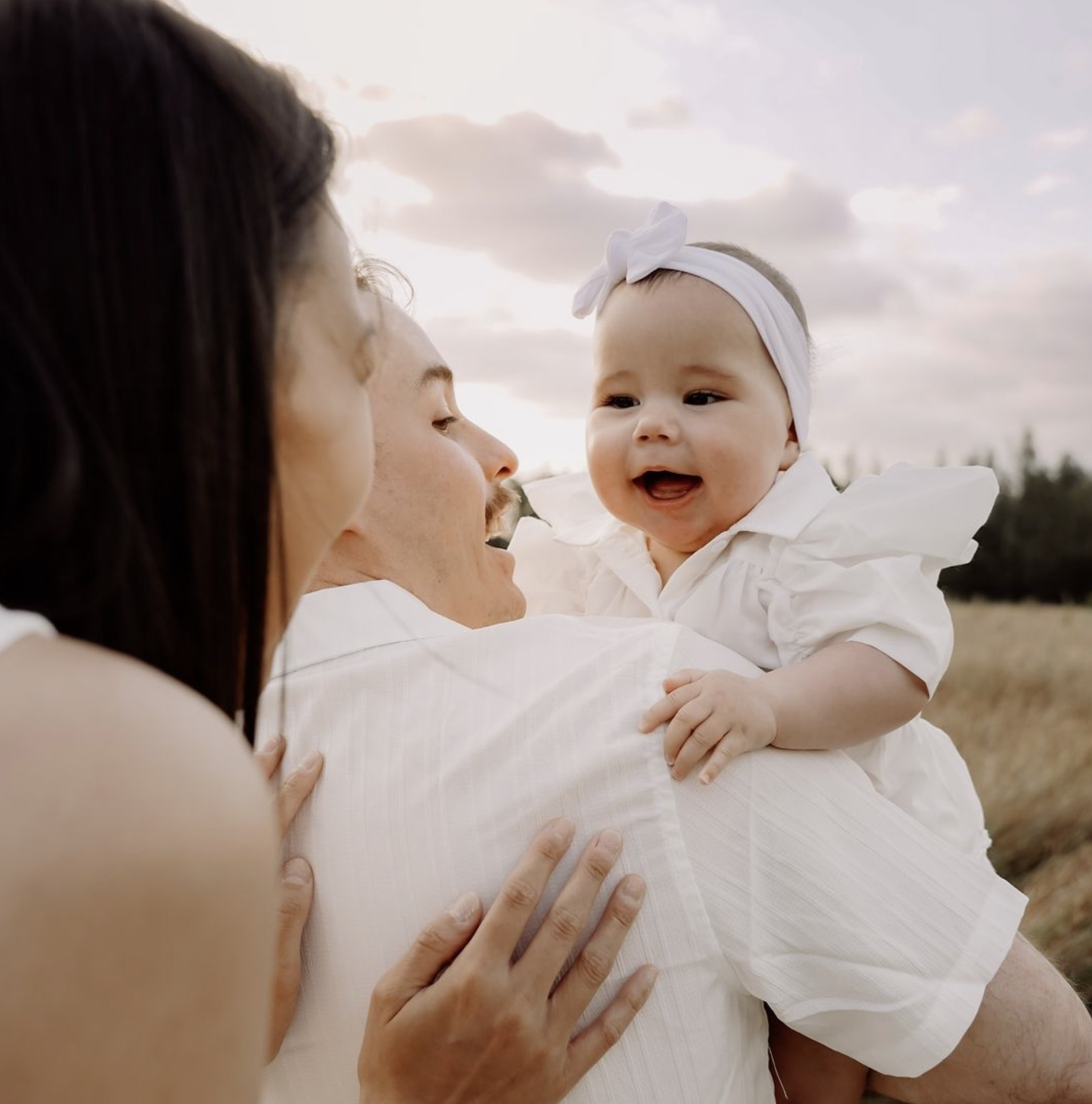 Several adults holding a smiling baby girl in an outdoor setting during sunset.