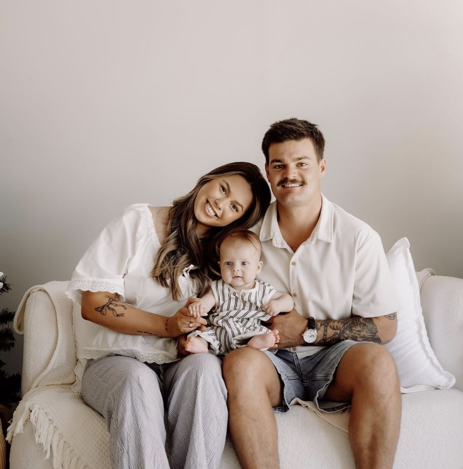 A family of four sitting on a white sofa, a woman with long wavy hair leaning her head on a man with a mustache and tattoos on his arm, holding a baby. The family is smiling in a cozy, neutral-colored living room.