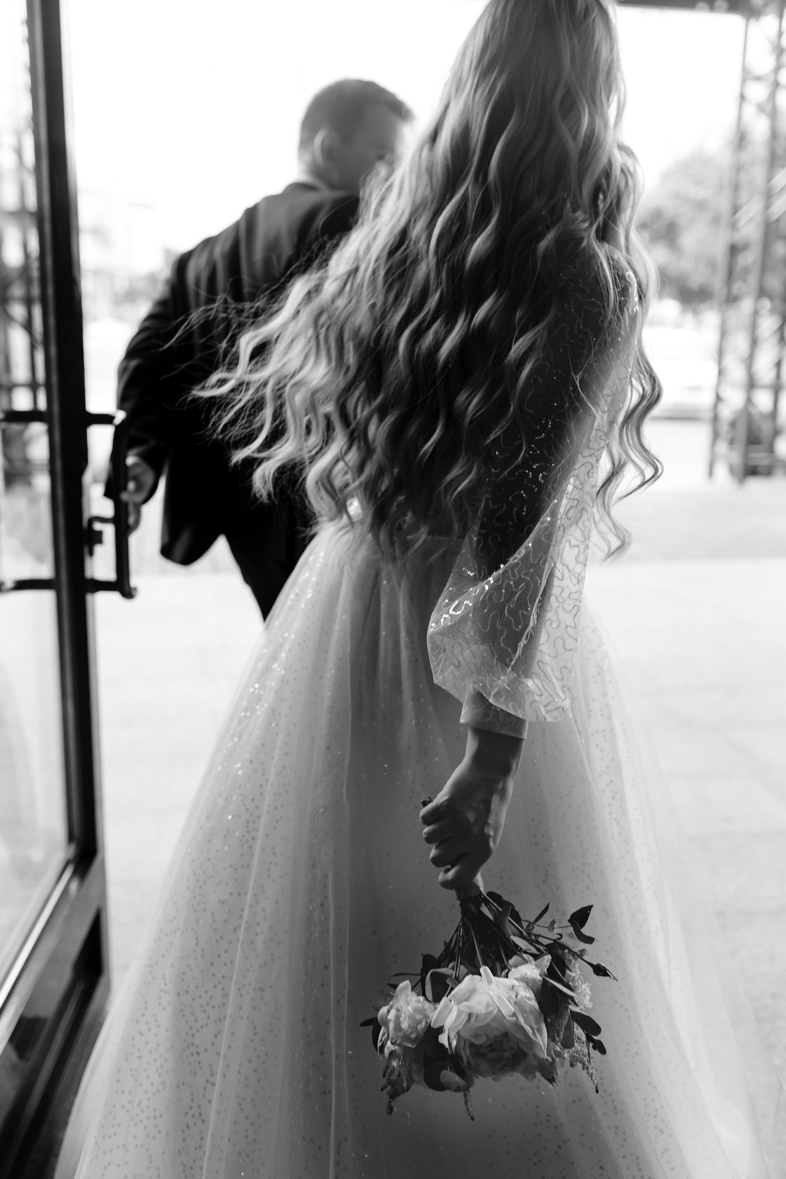 A young girl in a wedding dress holding a bouquet of flowers, standing near a window with a blurred background outside.
