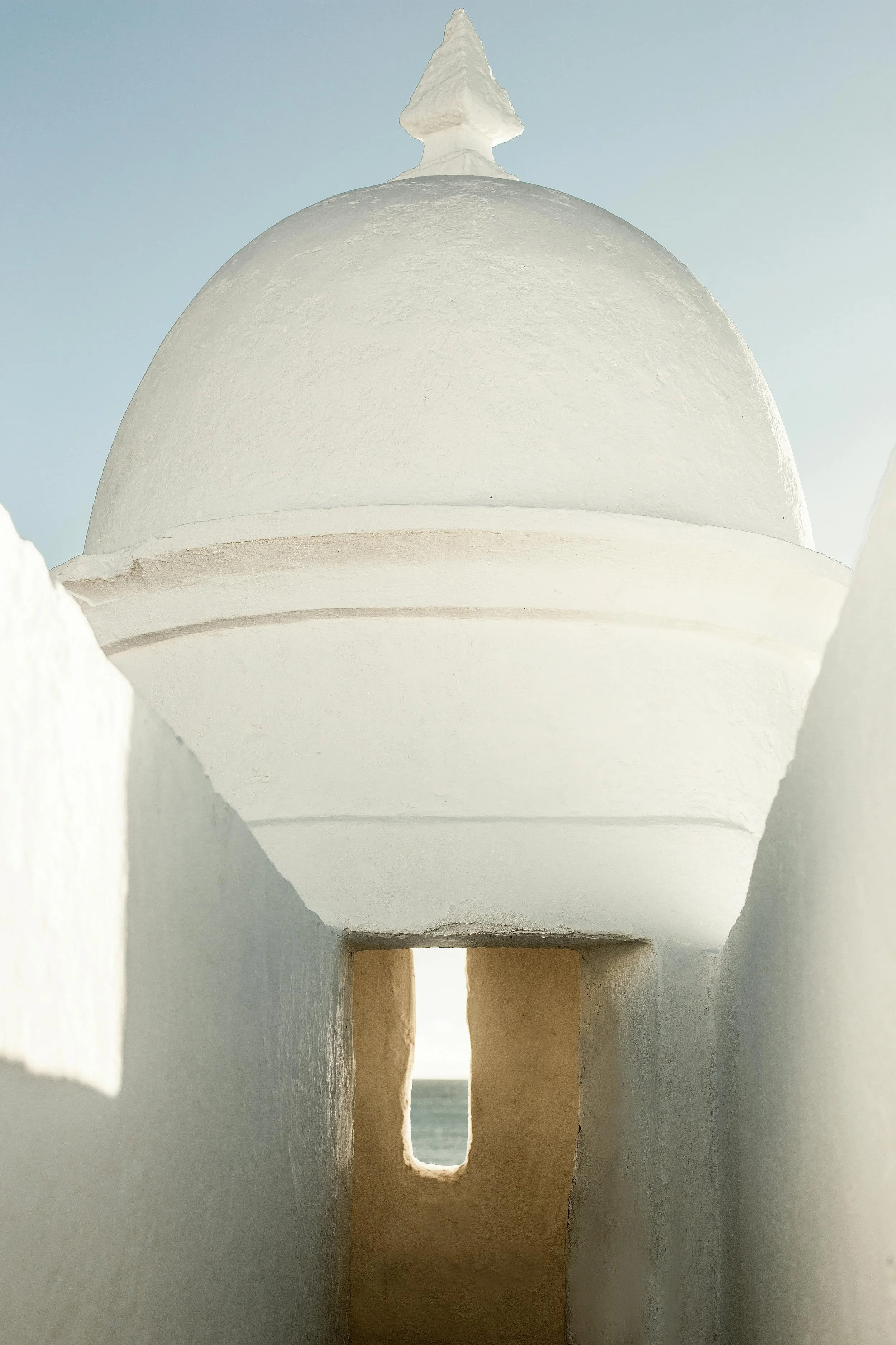 A white architectural structure with a domed top and a pointed spire, viewed from inside a corridor with openings and pillars, facing towards the sky and the sea.