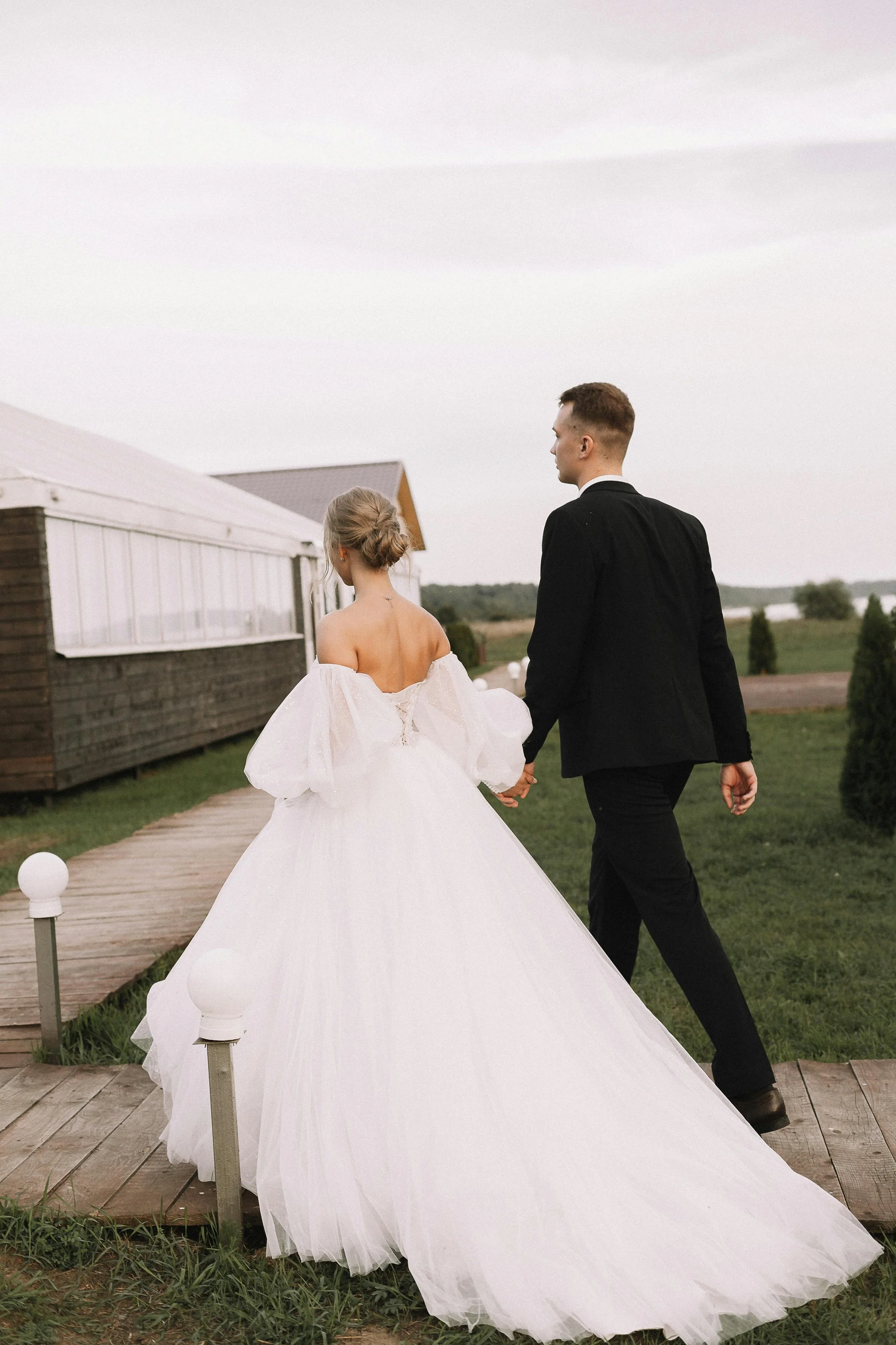 A bride and groom walking hand in hand on a wooden pathway outdoors, with the bride in a white wedding gown and the groom in a black suit, on a cloudy day with a landscape background.