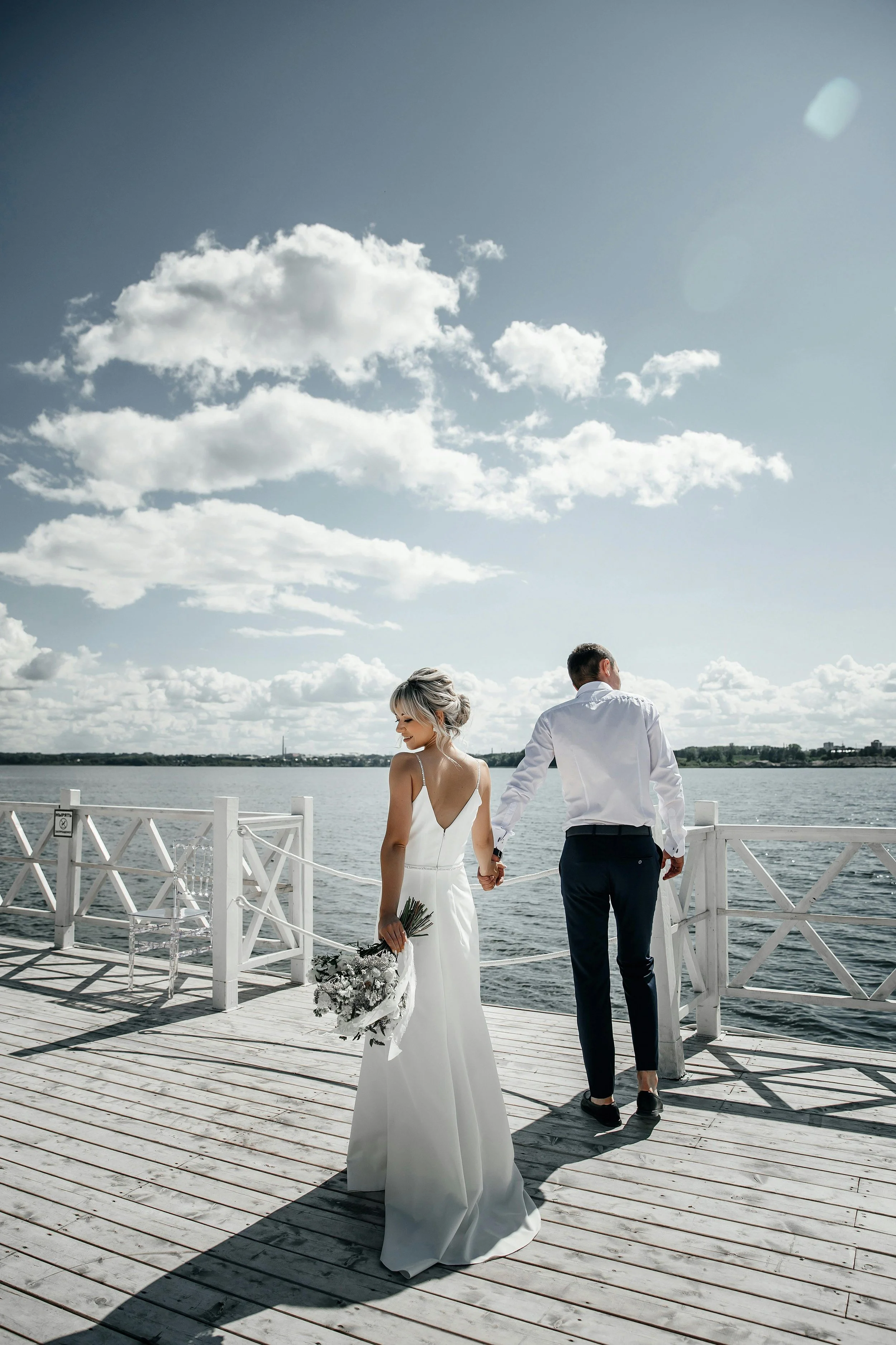 A bride in a white wedding dress holding a bouquet and a groom in a white shirt and dark pants holding hands, standing on a wooden dock by the water, with a cloudy sky above.