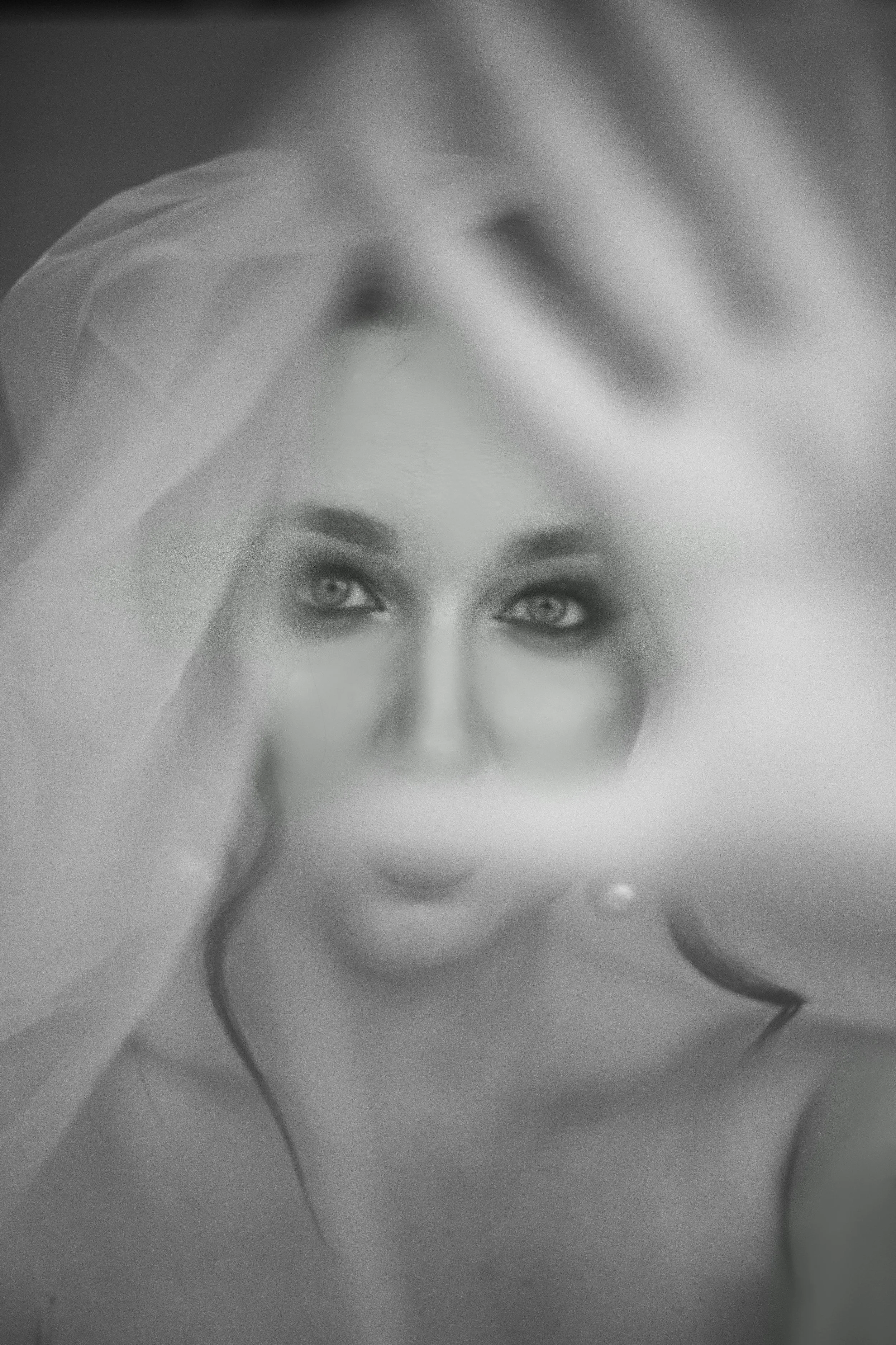 Close-up of a person's face with light-colored eyes and long, dark curly hair, holding a sheer fabric near their face.