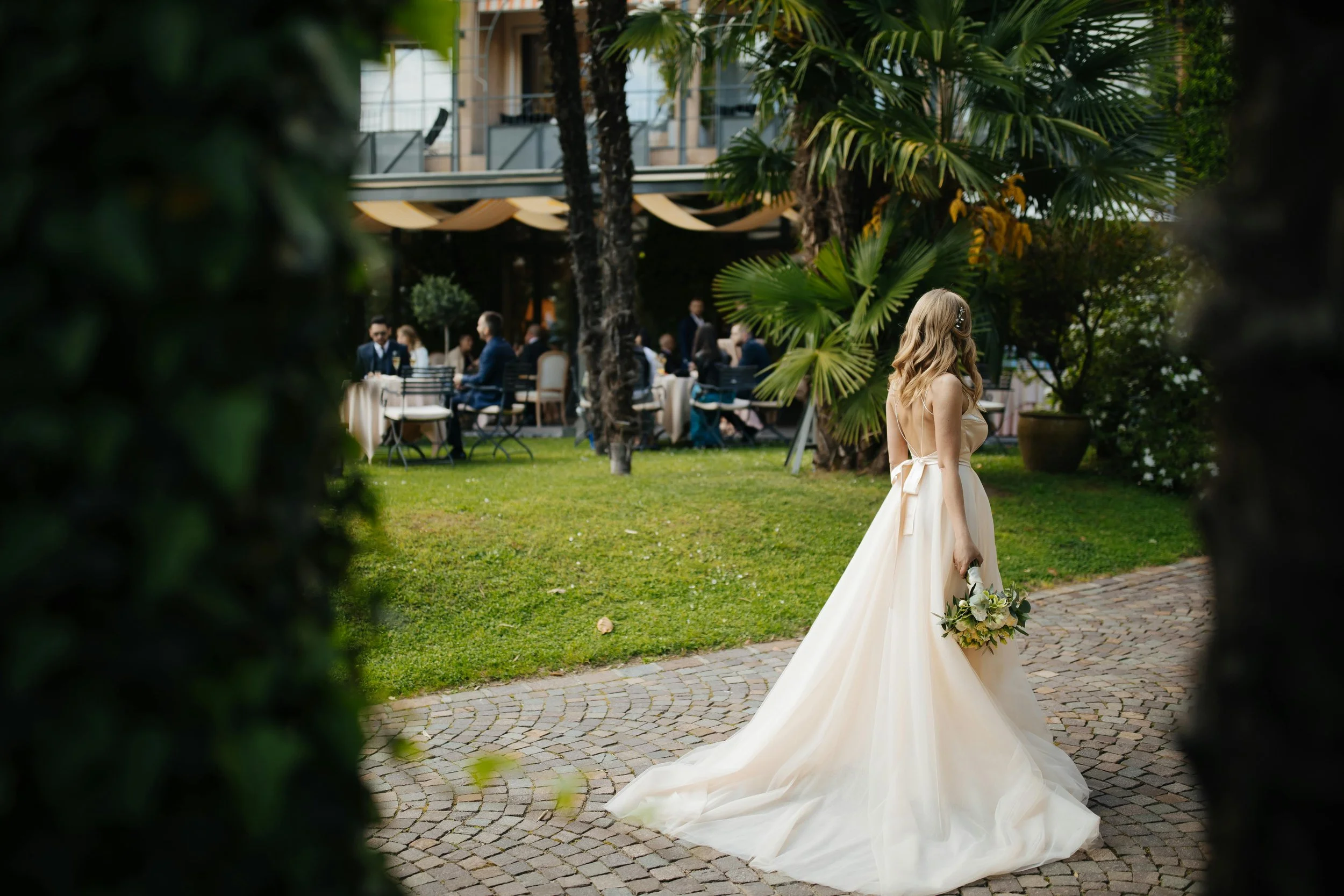 A bride in a white wedding gown standing outdoors on a cobblestone path holding a bouquet, with wedding guests dining at tables in the background surrounded by green trees and plants.