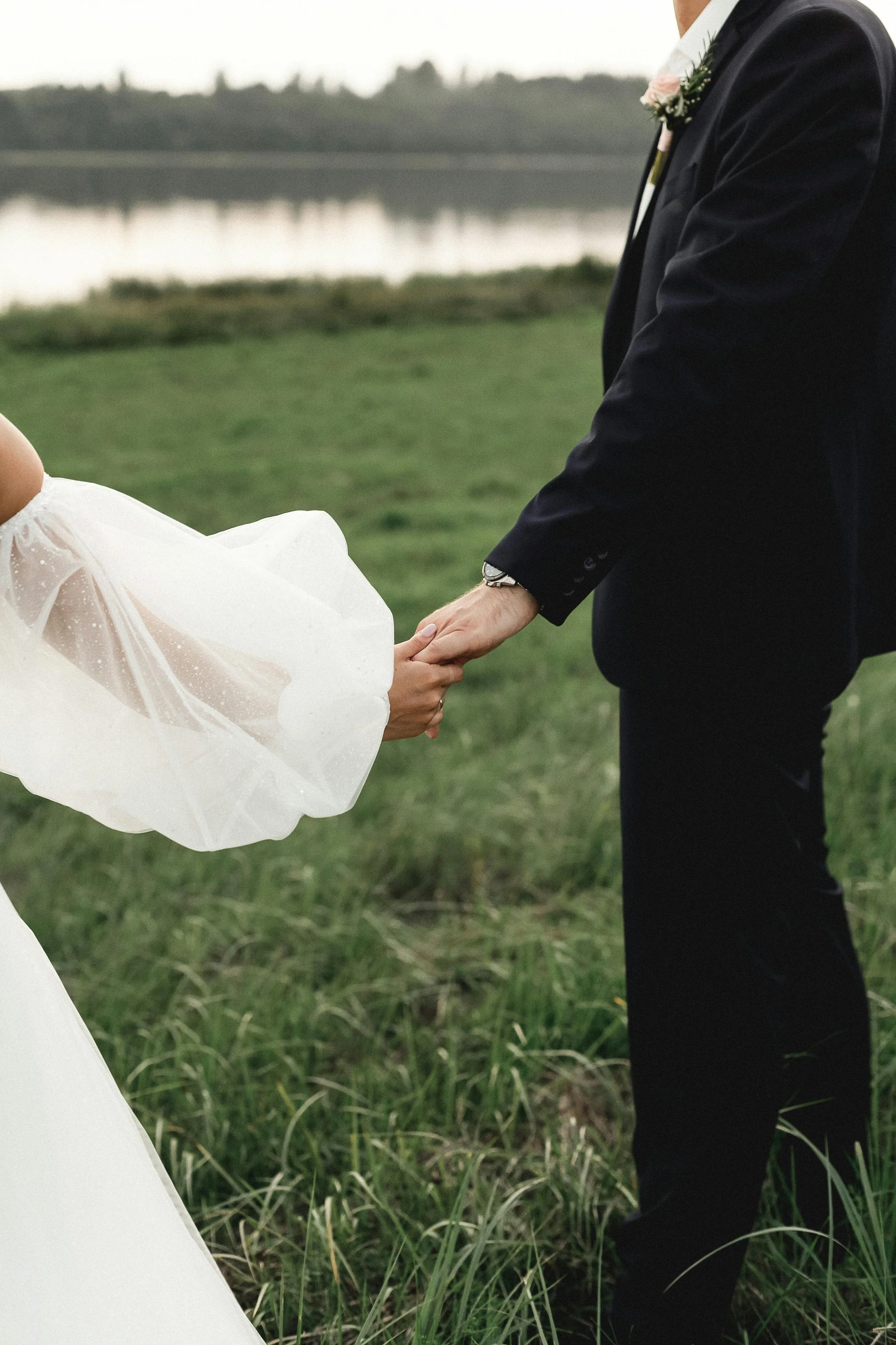 A bride and groom holding hands outdoors near a body of water, with grass and trees in the background.