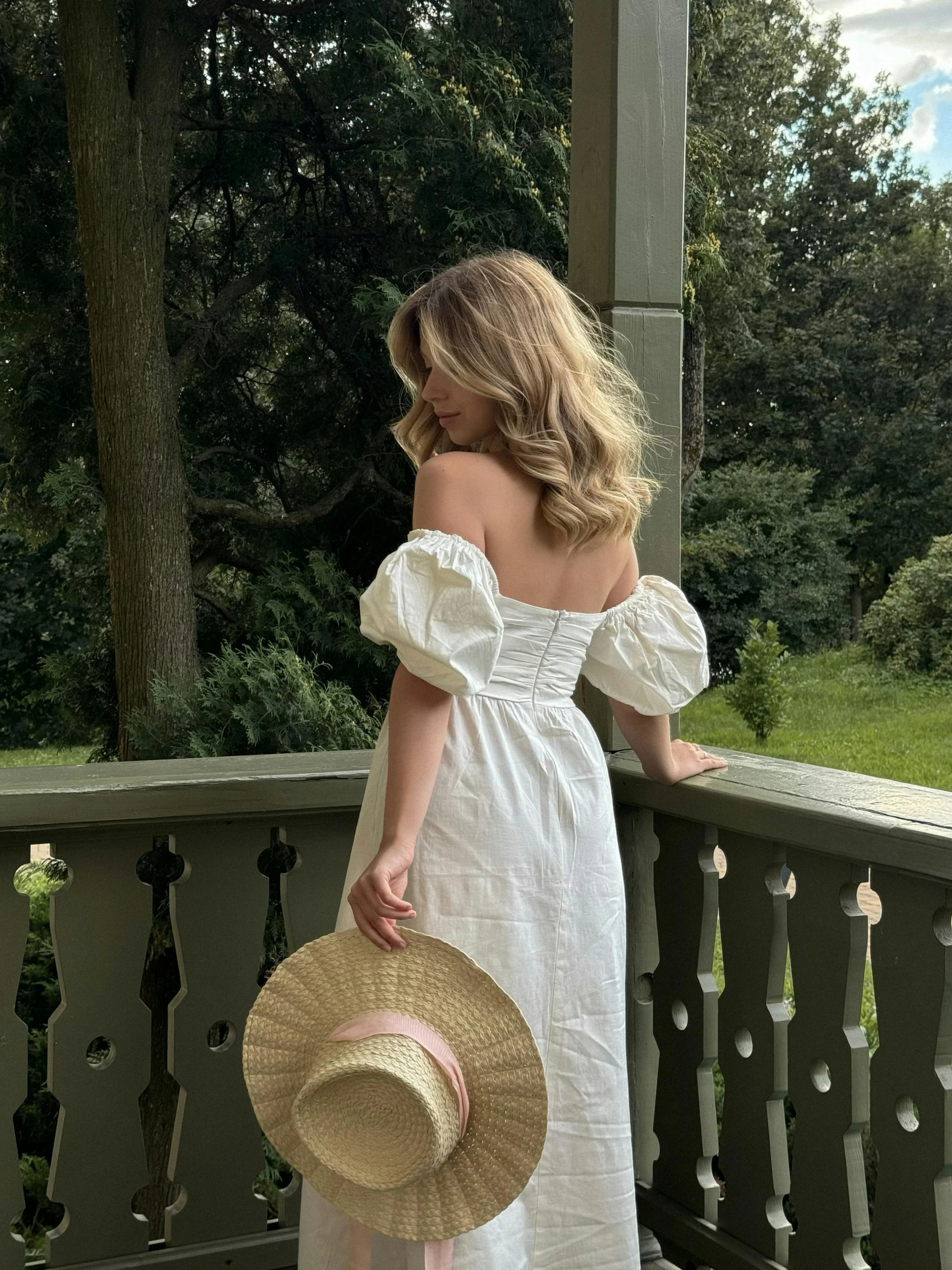 A woman with blonde hair wearing a white off-the-shoulder dress standing on a porch with a wooden railing, holding a straw hat, with trees and a partly cloudy sky in the background.
