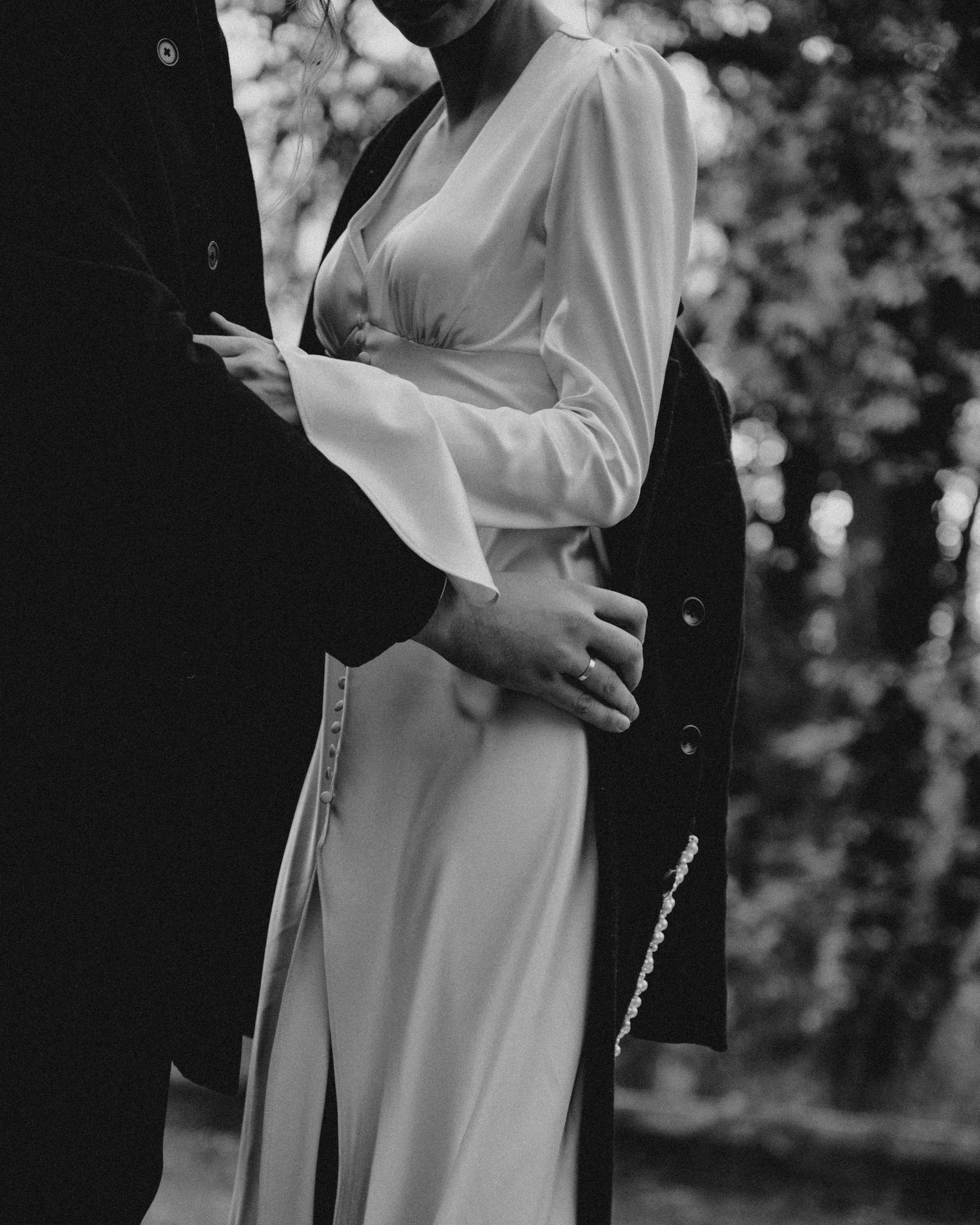 A black and white photo of a couple holding each other closely. The woman is wearing a light-colored dress with puffed sleeves and buttons on the side, and the man is dressed in a dark jacket with buttons. They are outdoors with blurred trees in the background.