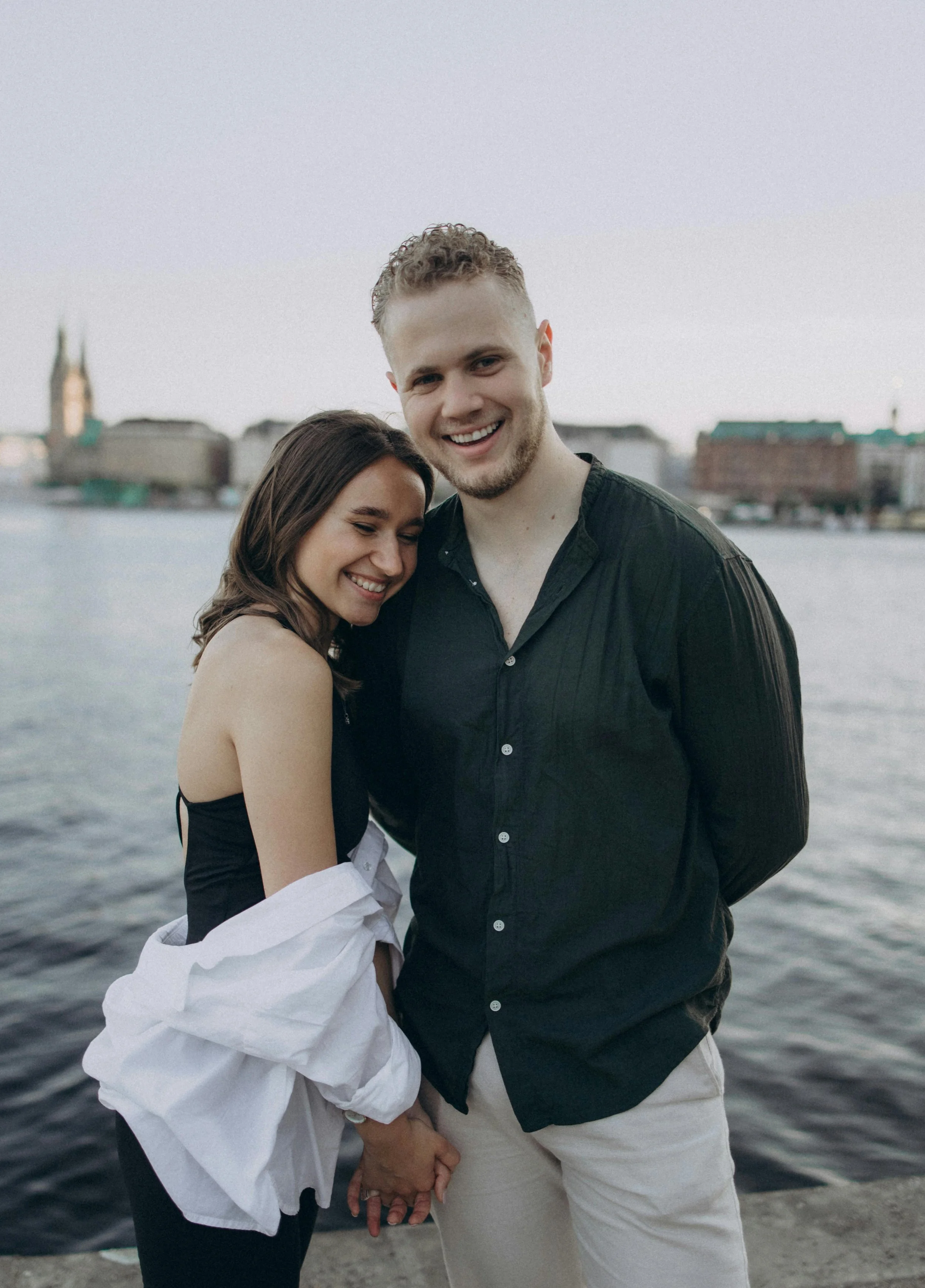 A smiling couple holding hands by the water, with cityscape buildings in the background.