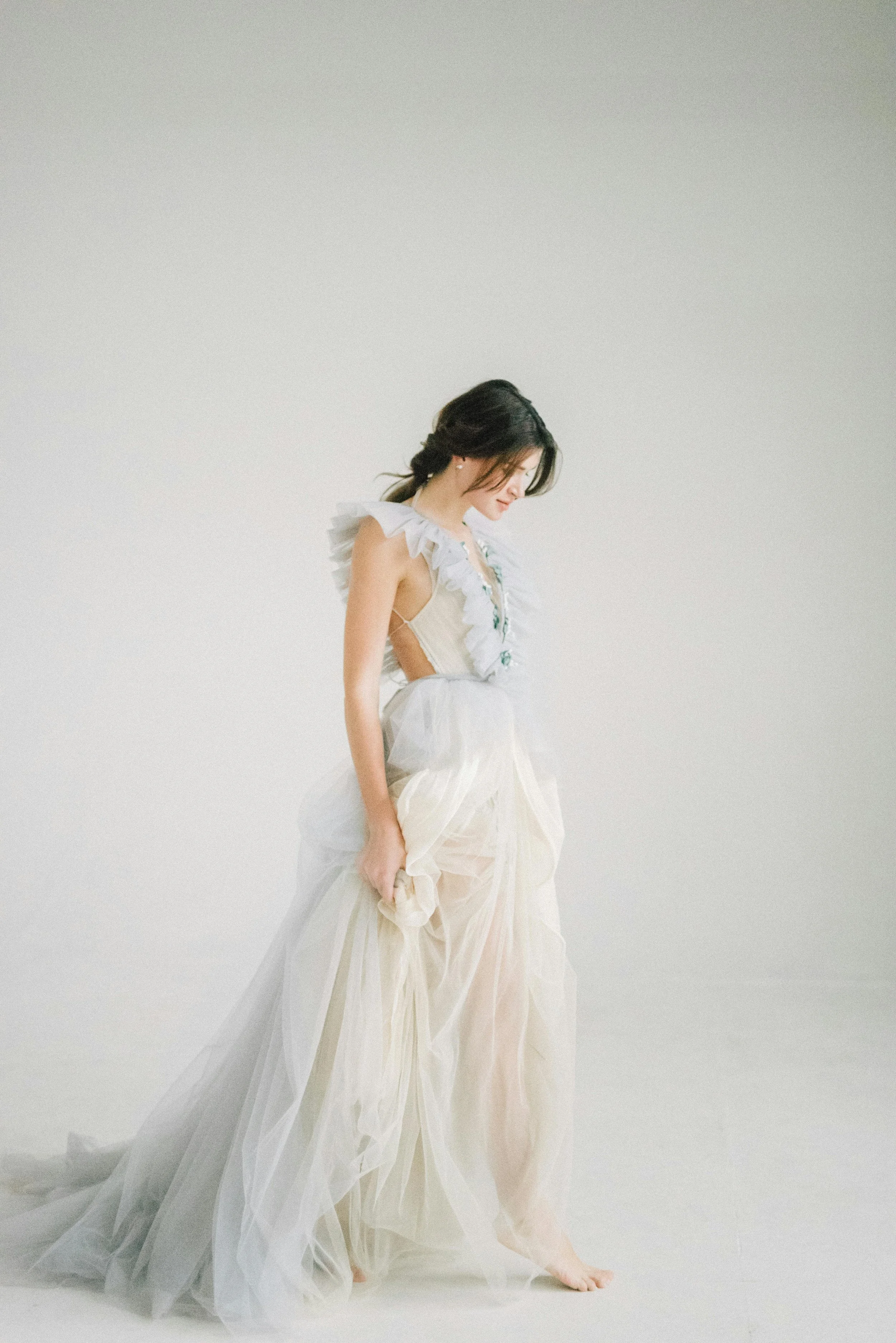 A woman with long dark hair in a loose ponytail wearing a white dress with ruffled details stands softly against a plain white background, looking down with a gentle expression.