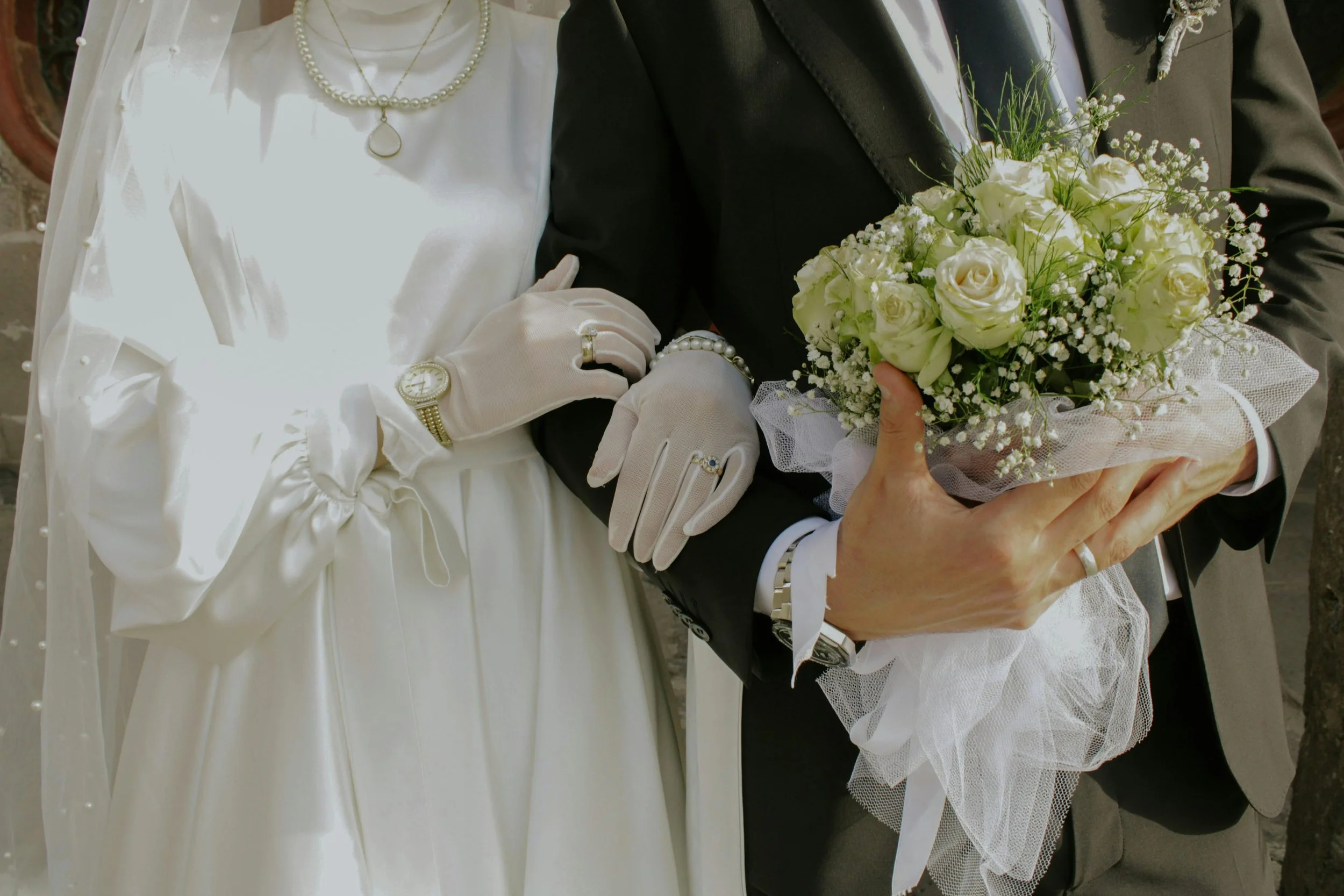 Close-up of a bride and groom holding a bouquet of white flowers during a wedding. The bride is dressed in a white gown and pearl necklace, and the groom is in a black suit. The bride's hand displays a gold watch and ring, and the groom's hand is on her waist.