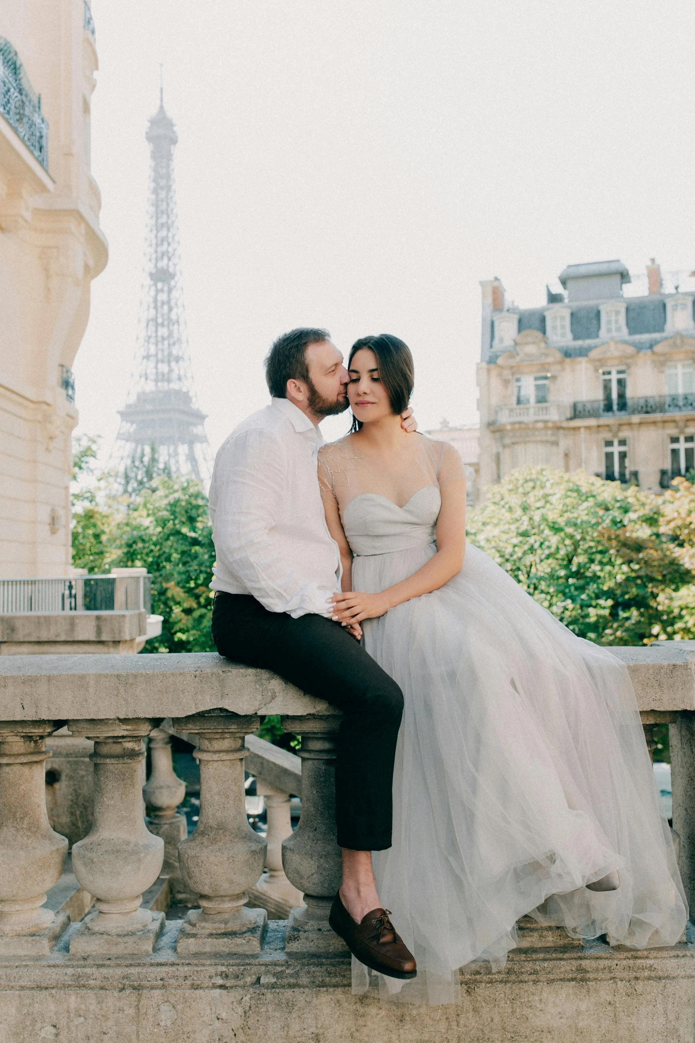 A couple dressed in wedding attire sitting on a stone balcony with the Eiffel Tower in the background. The man is wearing a white shirt and black pants, and the woman is wearing a white wedding dress.