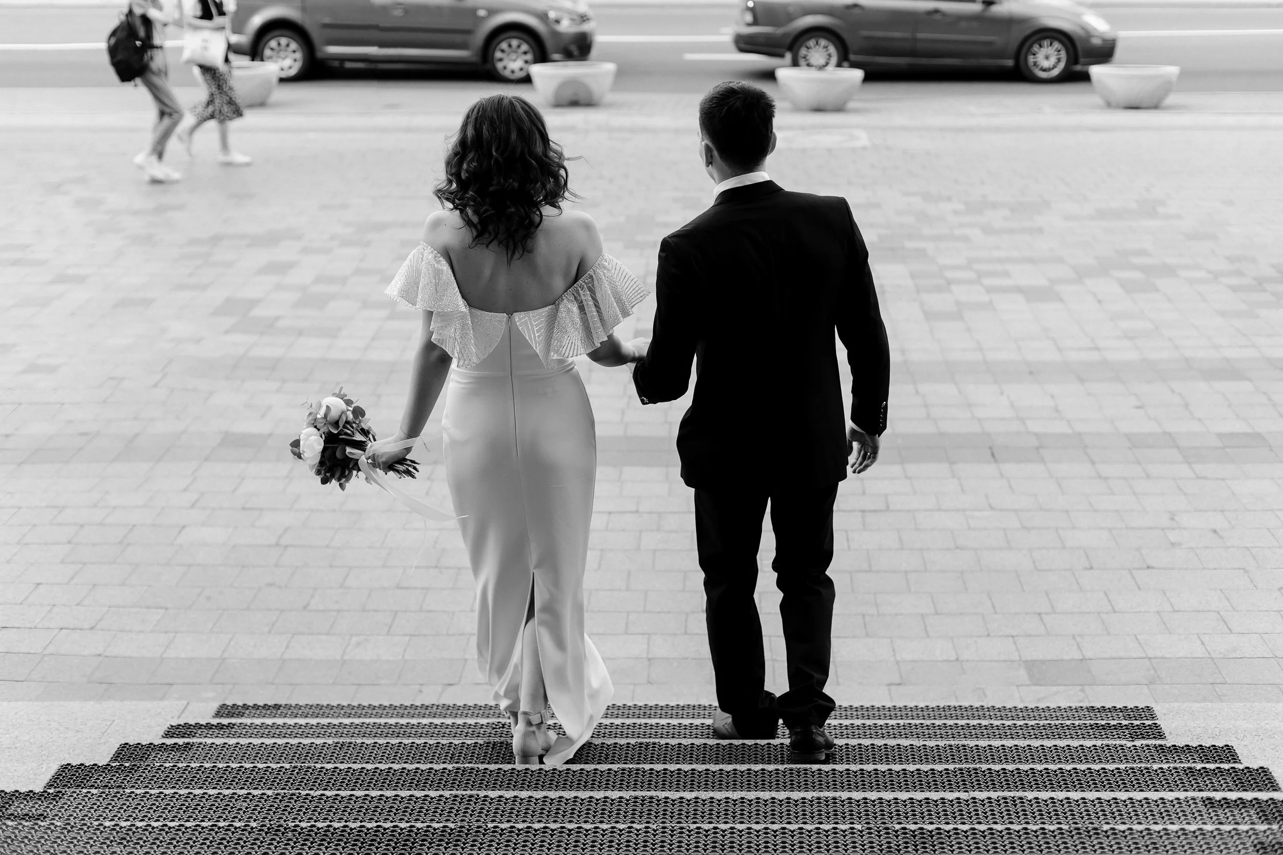 A couple dressed in wedding attire walking hand in hand on a city sidewalk, with cars passing by in the background.