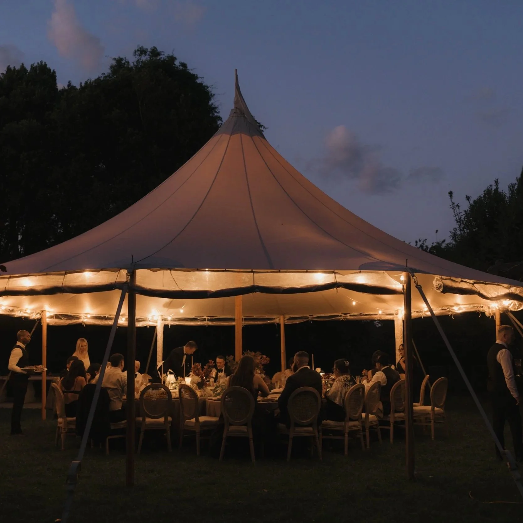 A large outdoor event tent decorated with string lights at dusk, with people seated at round tables inside, and some waiters serving food.