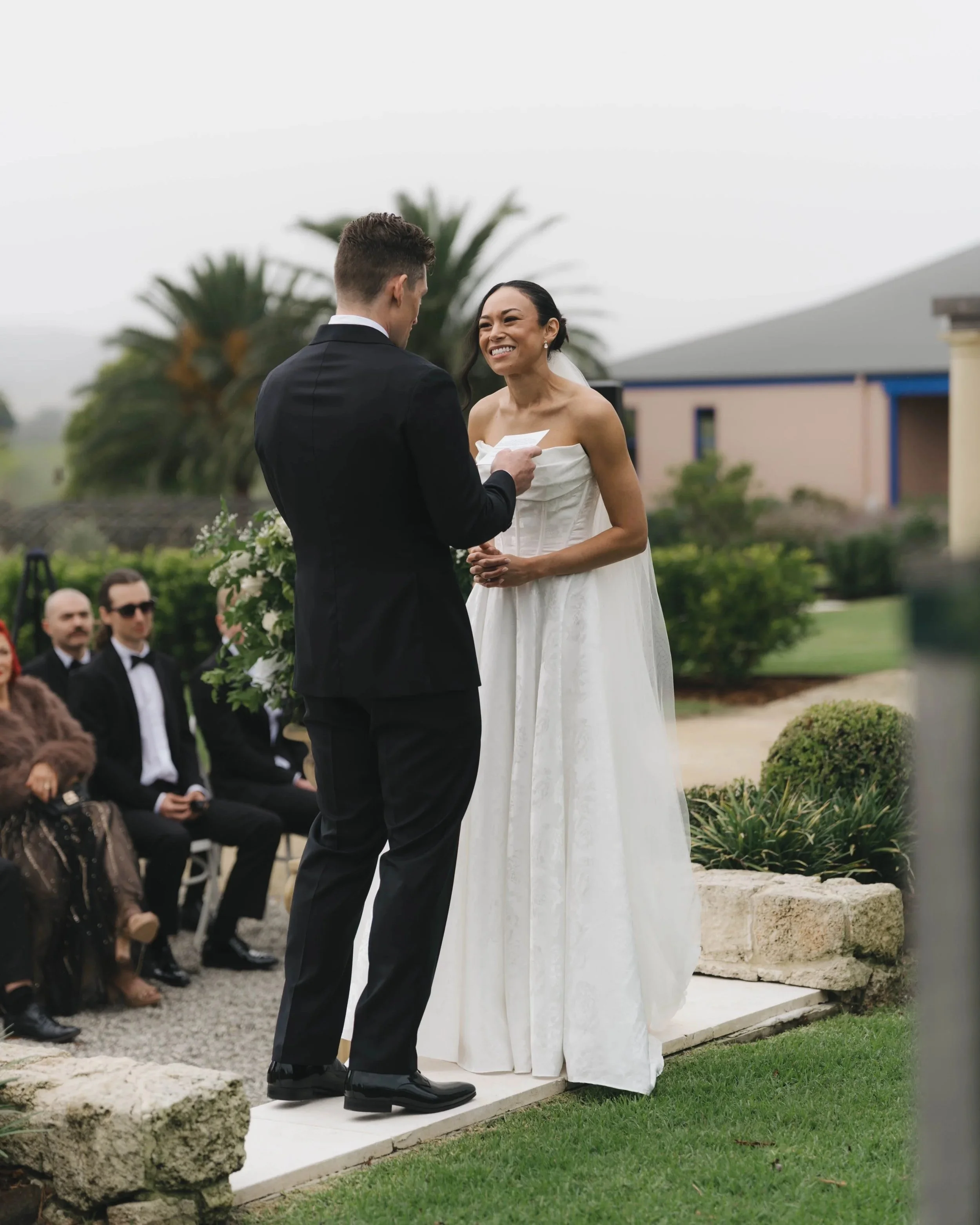 A couple getting married outdoors, the groom reading vows from a small paper, the bride smiling and looking at him. Guests seated nearby, with buildings, trees, and a cloudy sky in the background.
