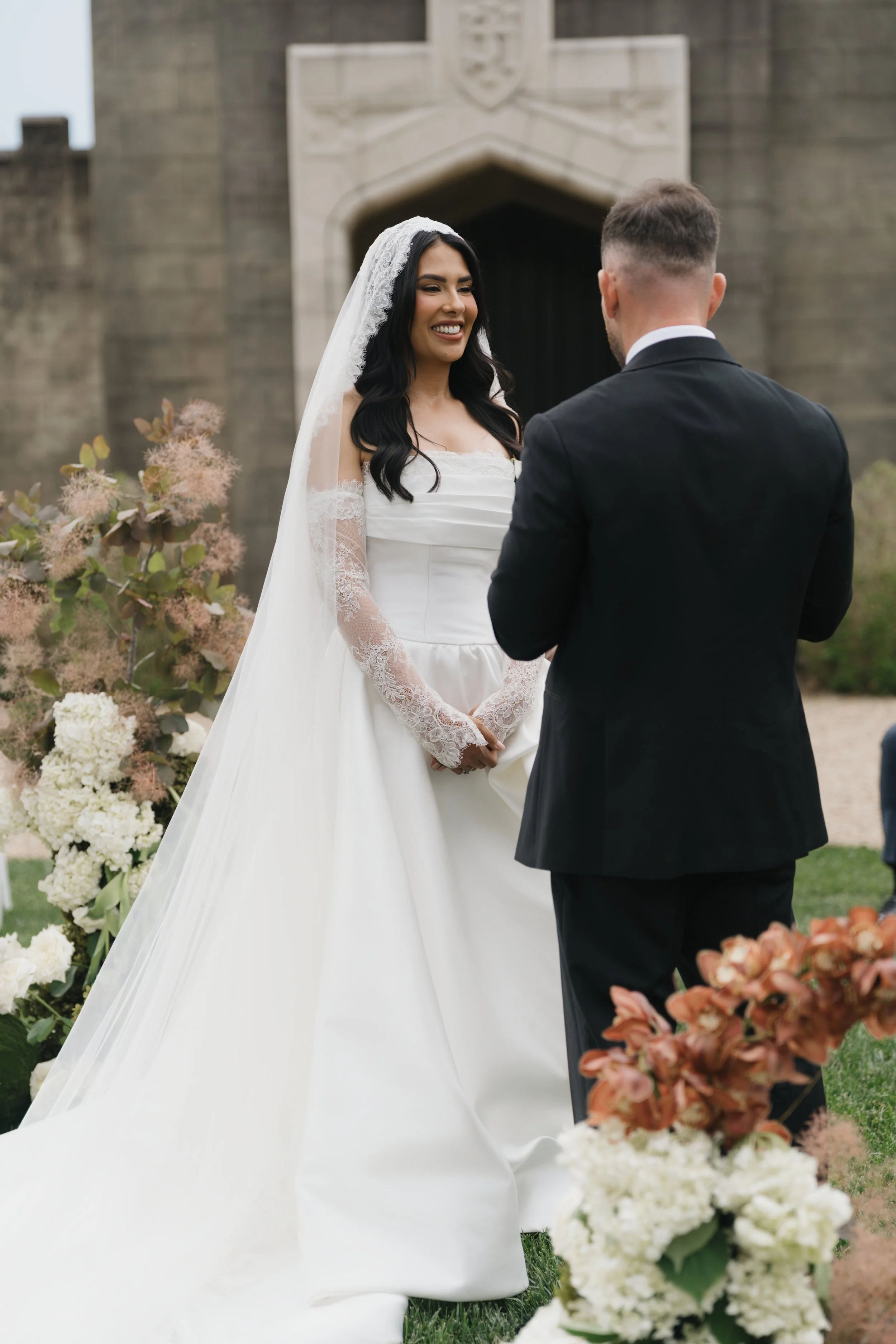A bride and groom exchanging vows at an outdoor wedding, with the bride wearing a white wedding gown and veil, and the groom in a black tuxedo, surrounded by floral decorations.
