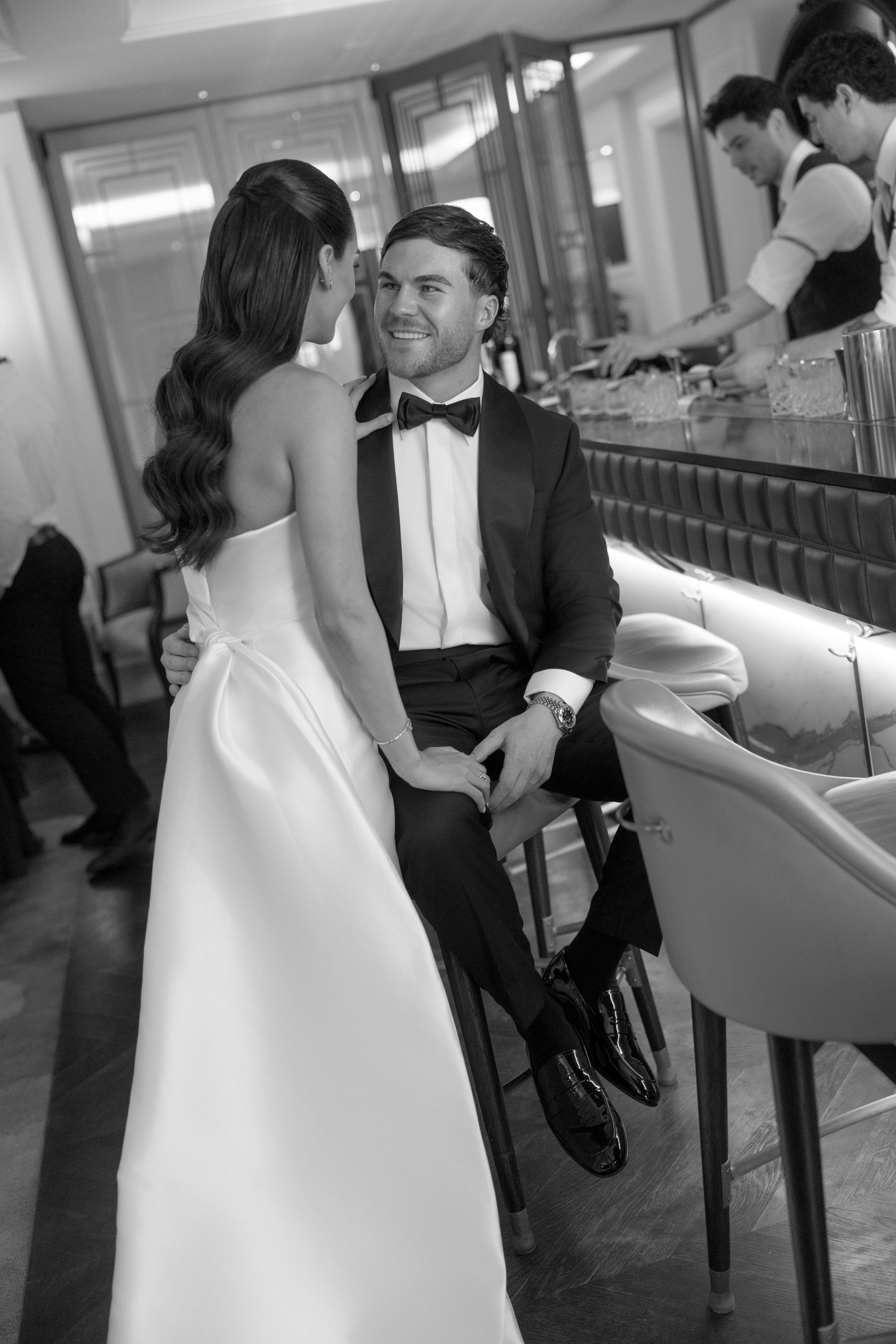 A man in a tuxedo and woman in a wedding dress sitting at a bar, sharing a moment. Two bartenders are behind the bar preparing drinks.