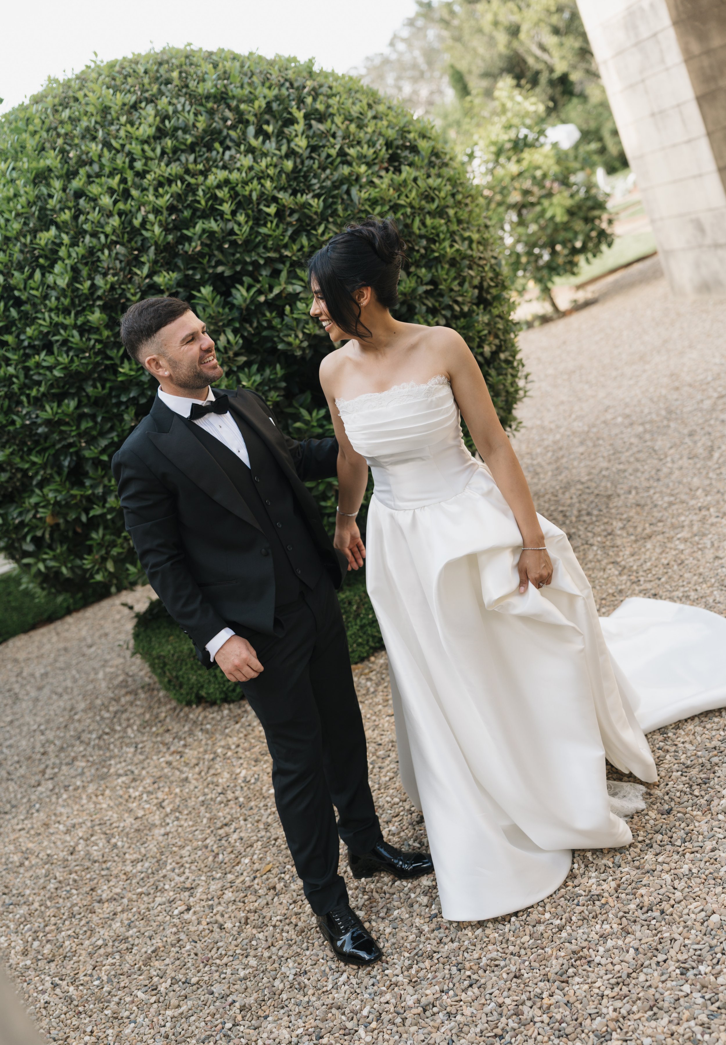 A bride and groom are smiling and holding hands outdoors near a large green bush, with the bride in a strapless white wedding dress and the groom in a black tuxedo.