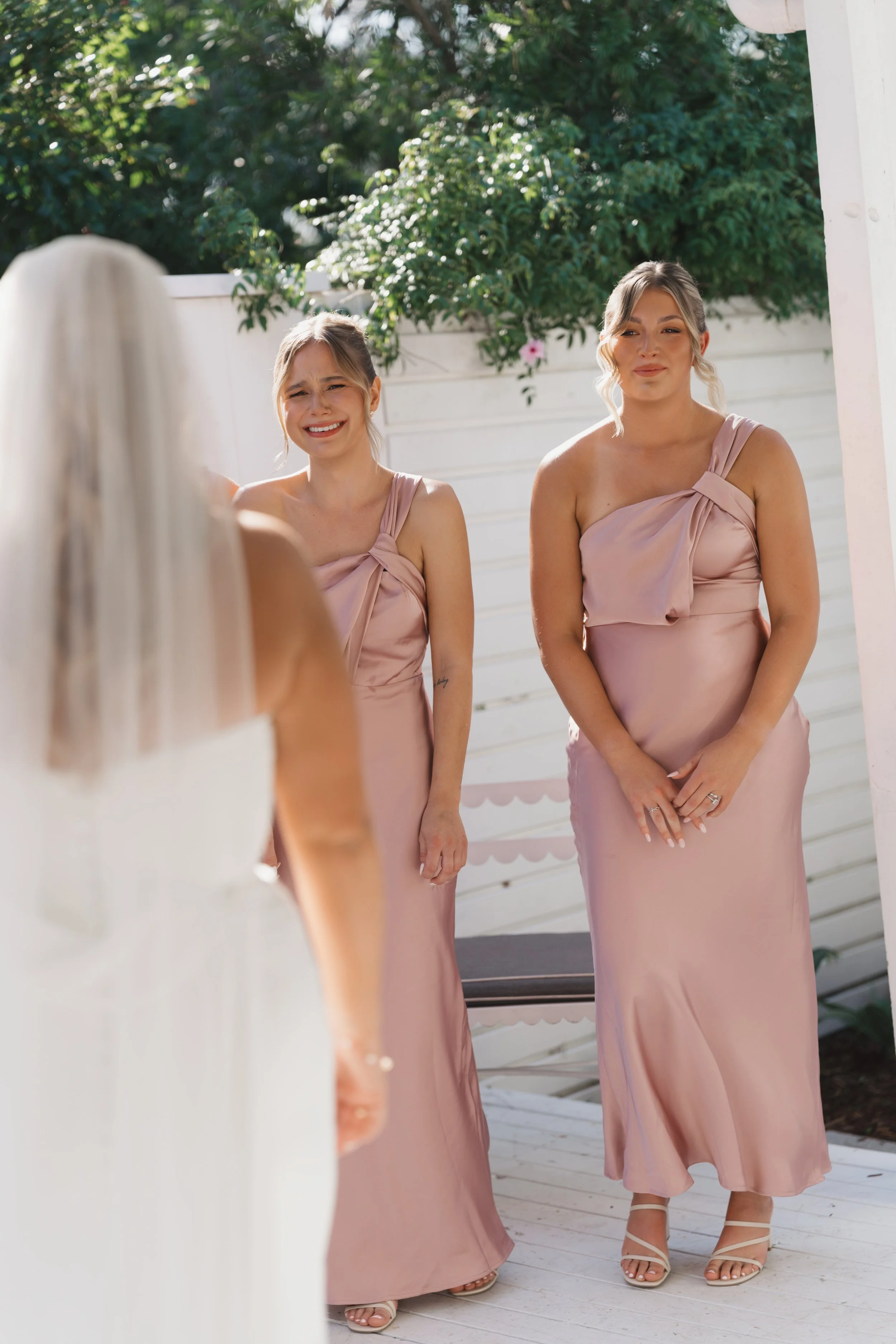 Three women in pink dresses standing outside on a white wooden deck, looking at a woman with long white hair and a veil, possibly at a wedding.