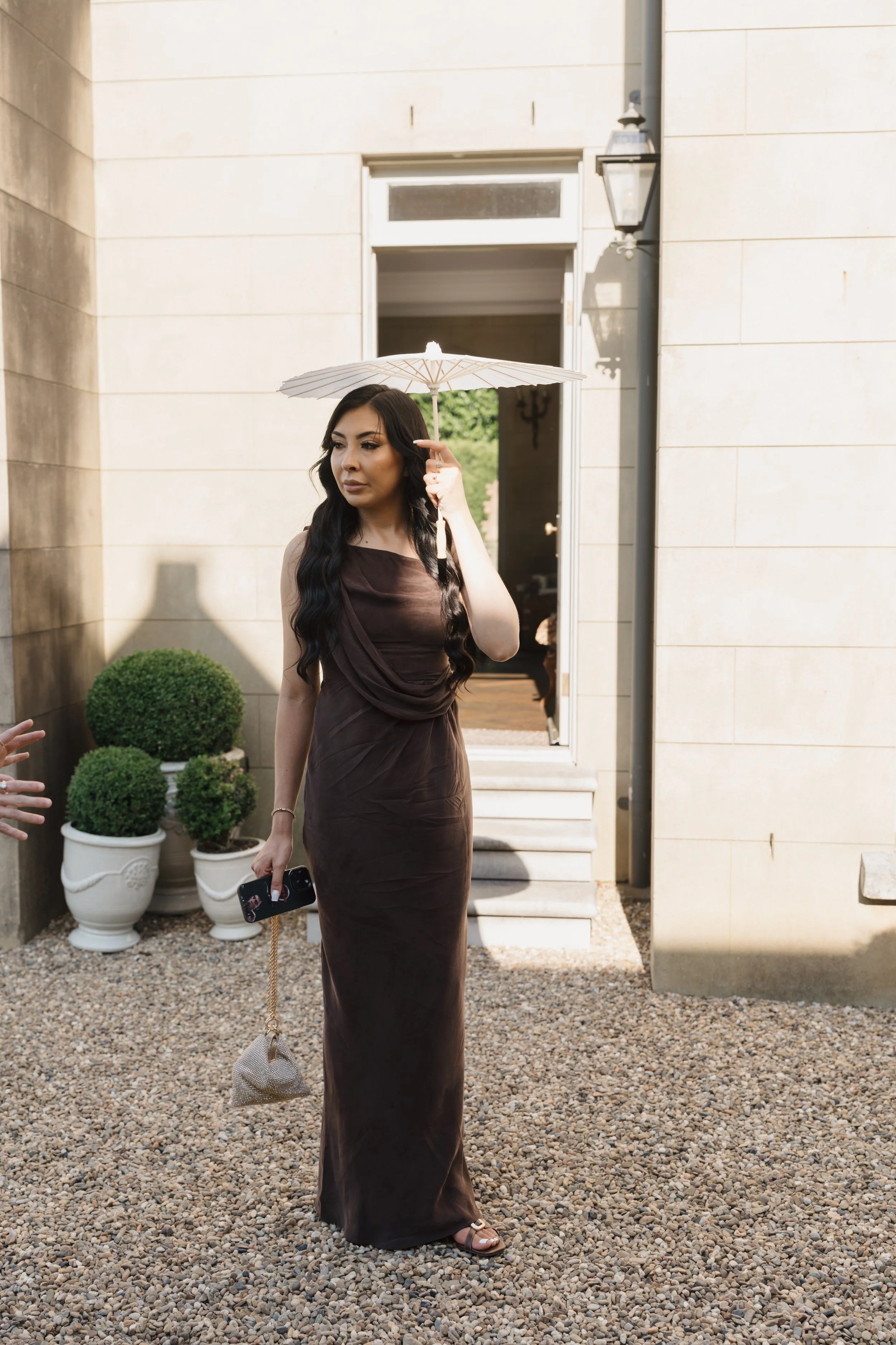 A woman with long black hair wearing a brown dress holding an umbrella and a Smartphone walking on a gravel path outside a beige building with potted plants.