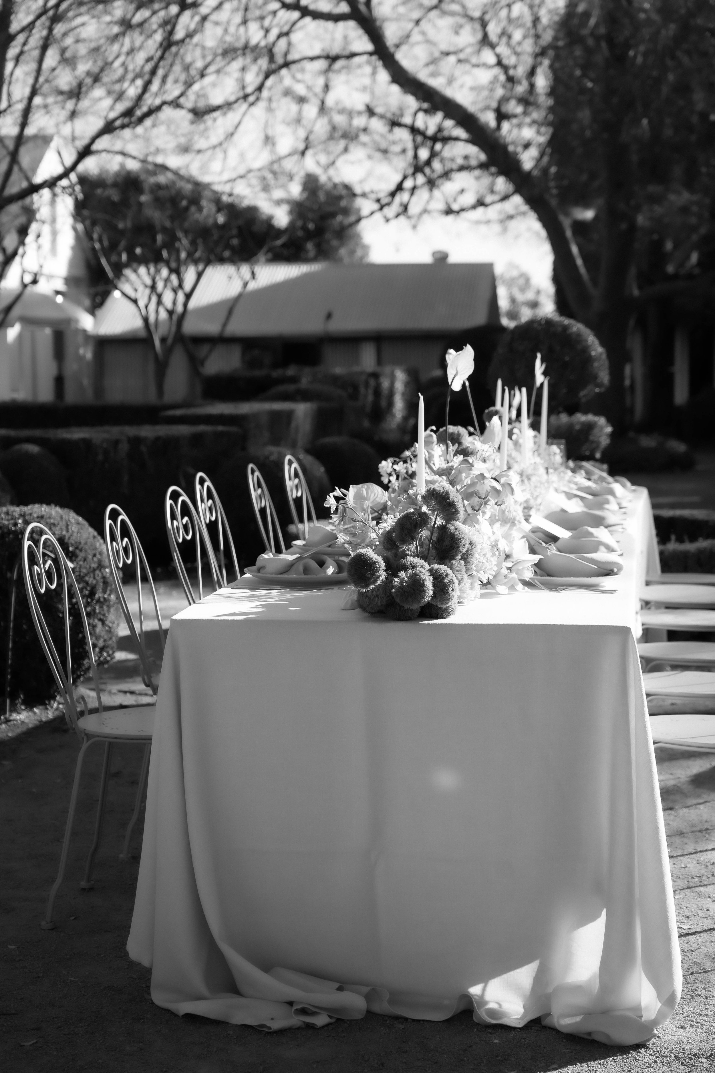 A long dining table with a white tablecloth decorated with floral arrangements and candles, outdoors near trees and a building with a sloped roof.