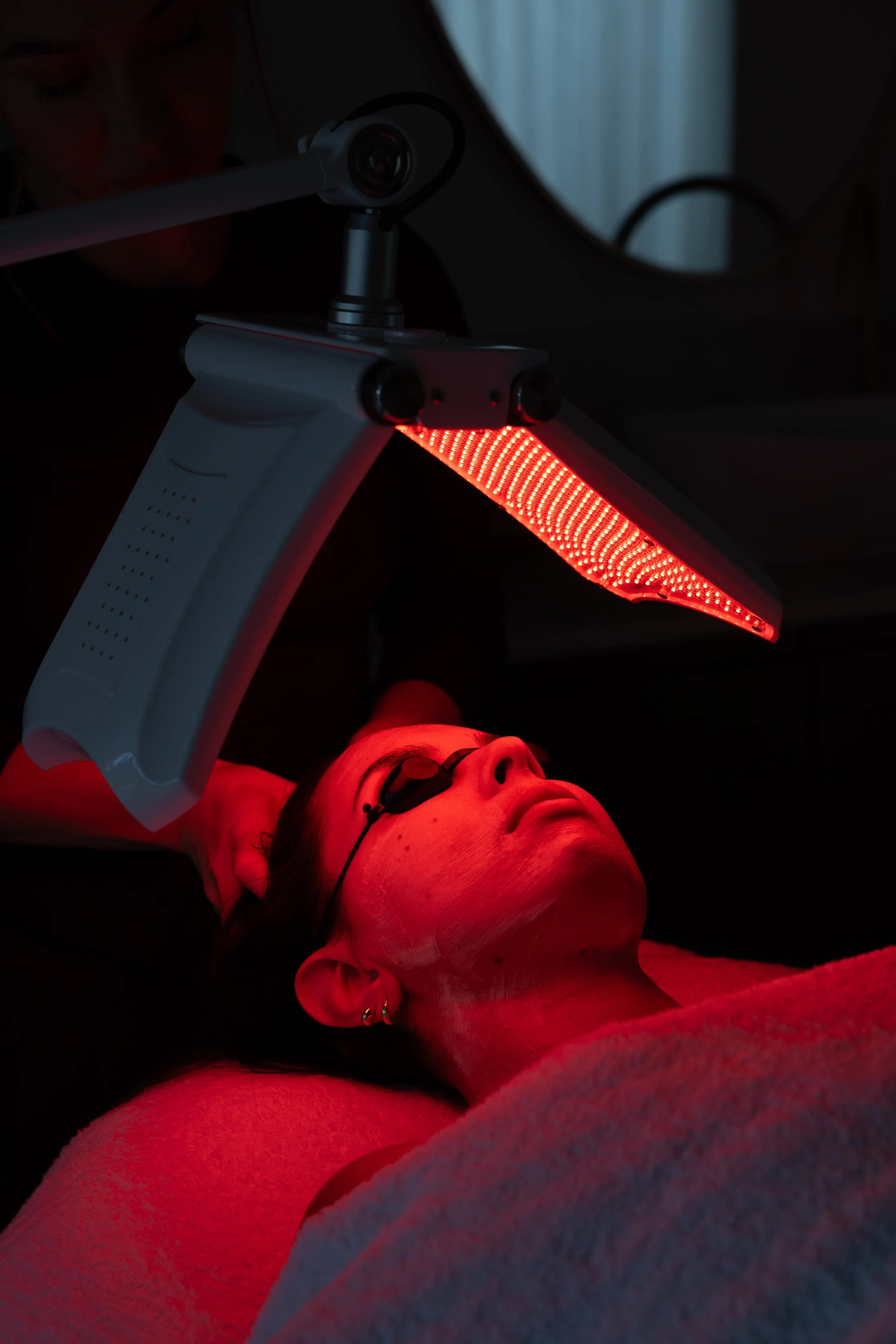 A woman lying down during a red light therapy session, with a red LED light panel shining on her face and chest.