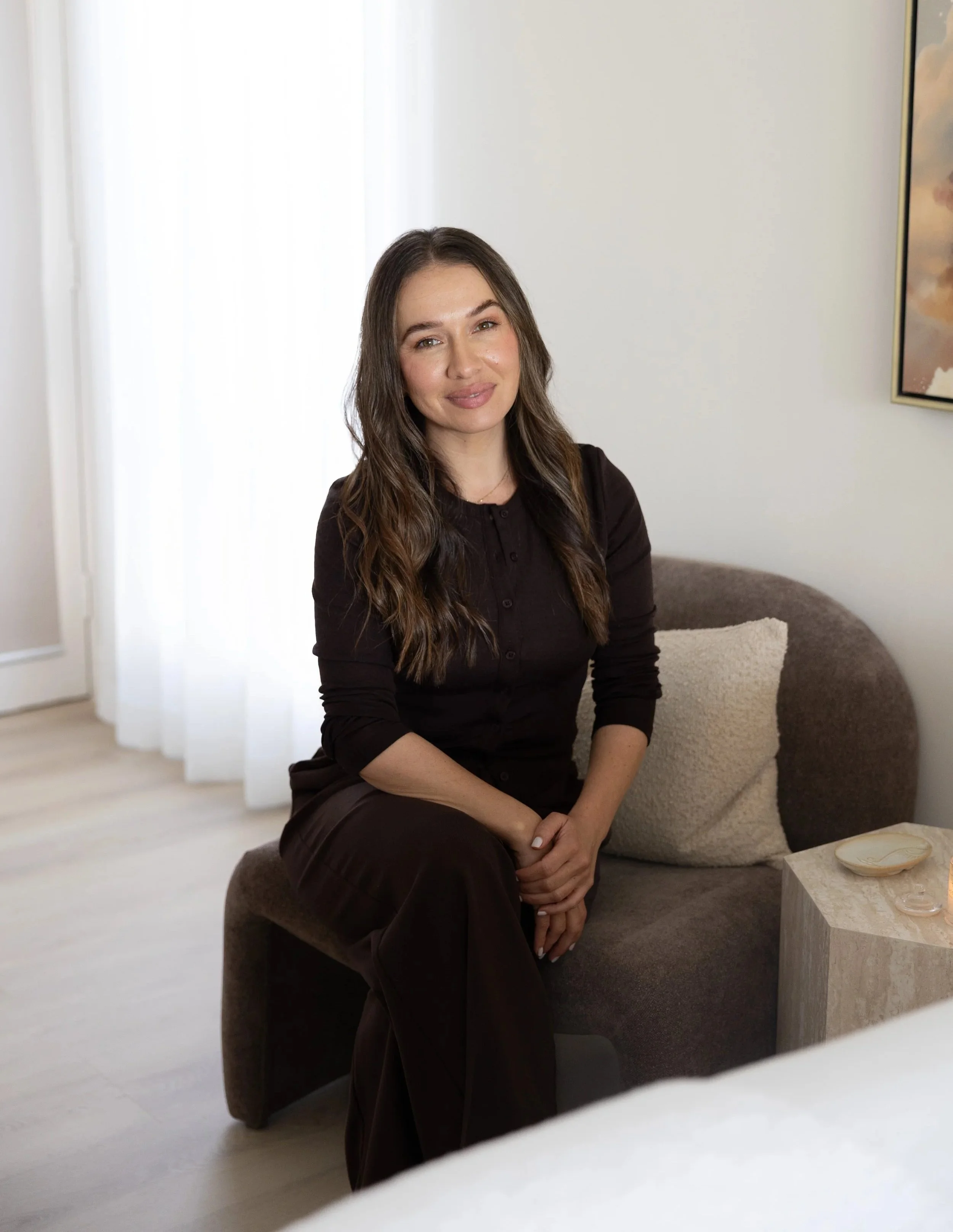 A woman with long brown hair, wearing a dark top, sitting on a brown sofa in a well-lit room with white walls. There is a framed picture on the wall and a small side table with a decorative dish.