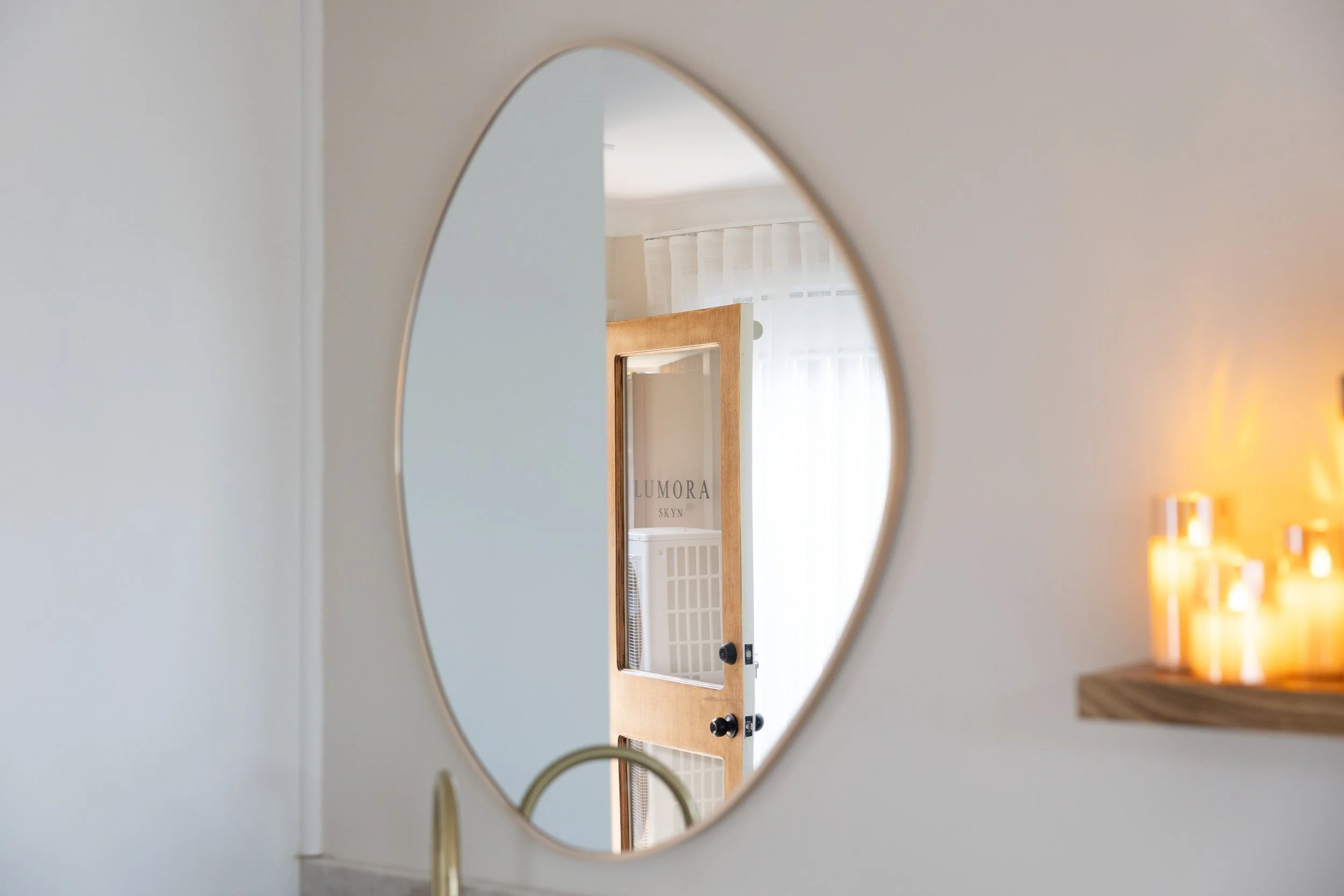 An oval mirror on a white wall reflecting a room with a framed artwork, a window with sheer curtains, and a dresser with candles.