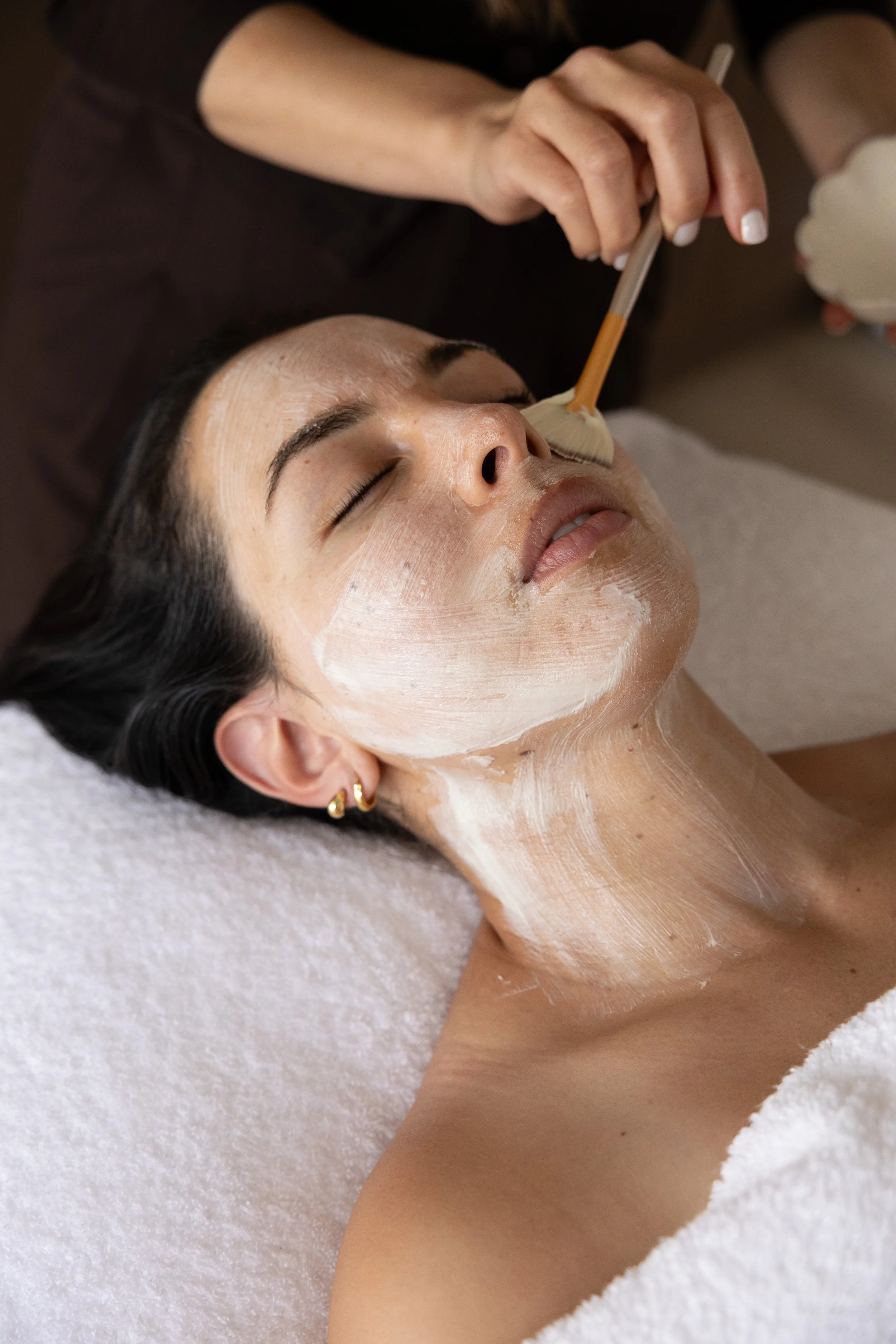 A woman lying on a white towel with closed eyes, receiving a facial treatment with a brush applying a cream or mask on her face.
