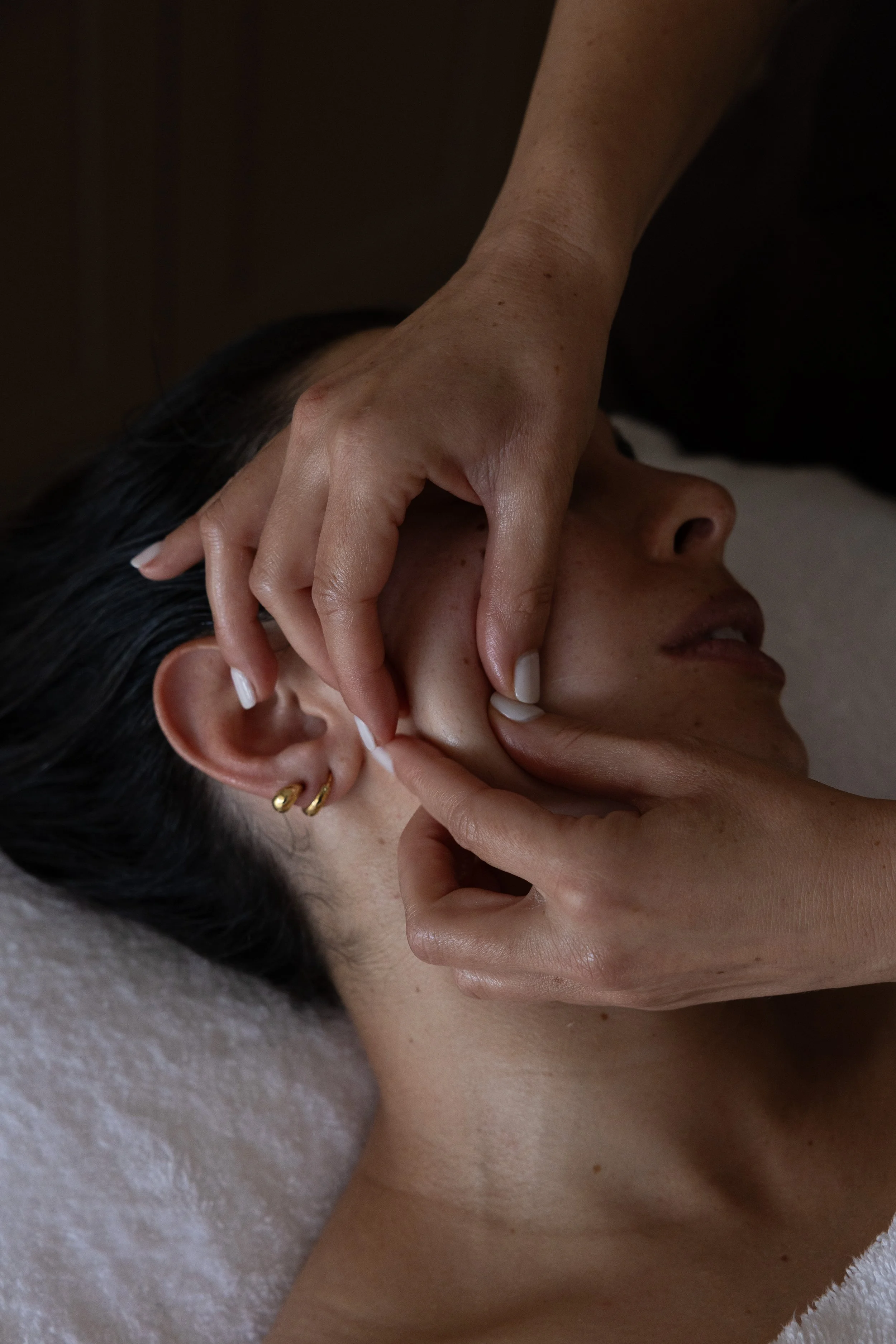 Person receiving or giving a massage or ear cleaning, close-up view of their head, hand on ear, dark hair, gold earring, lying on a soft, light-colored surface.