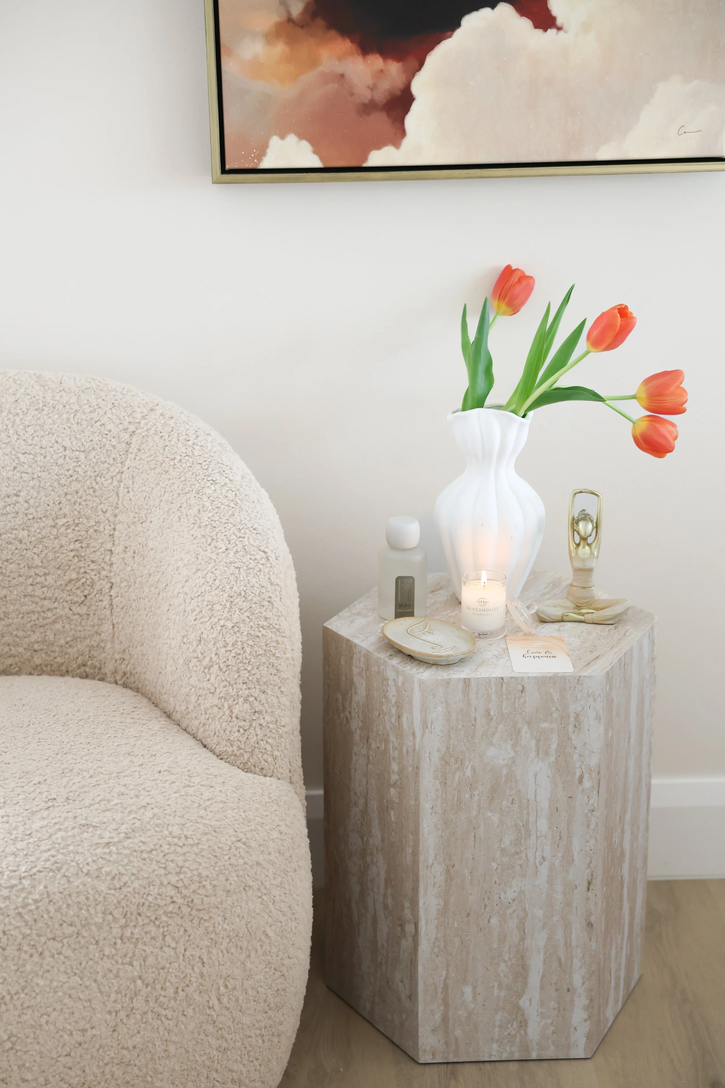 A cozy living space featuring a plush, beige armchair, a small marble side table, a white vase with orange tulips, a candle, and decorative items, all set against a plain white wall with a colorful abstract painting.